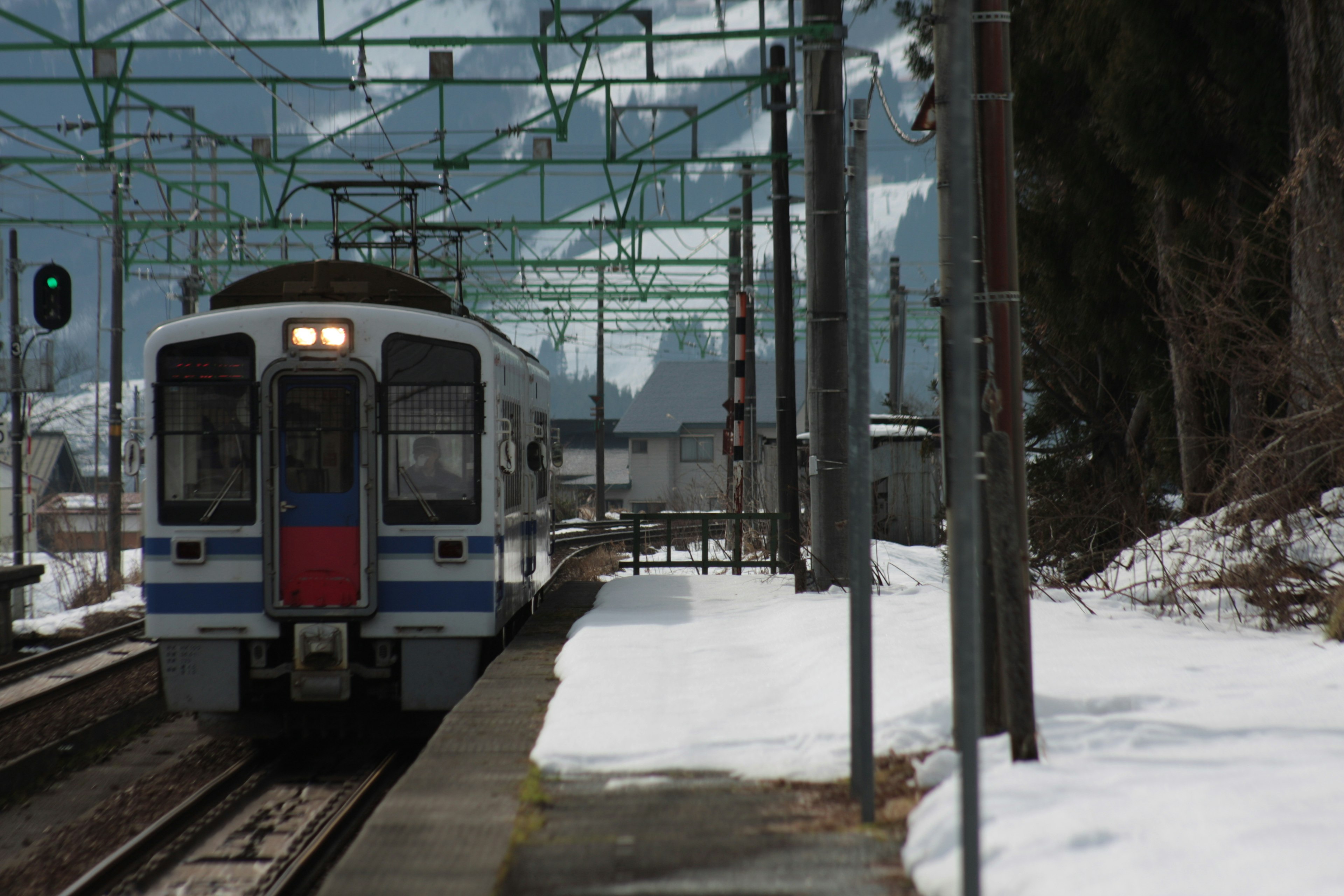 Train stopped at a snowy station green signal light and railway tracks nearby mountains