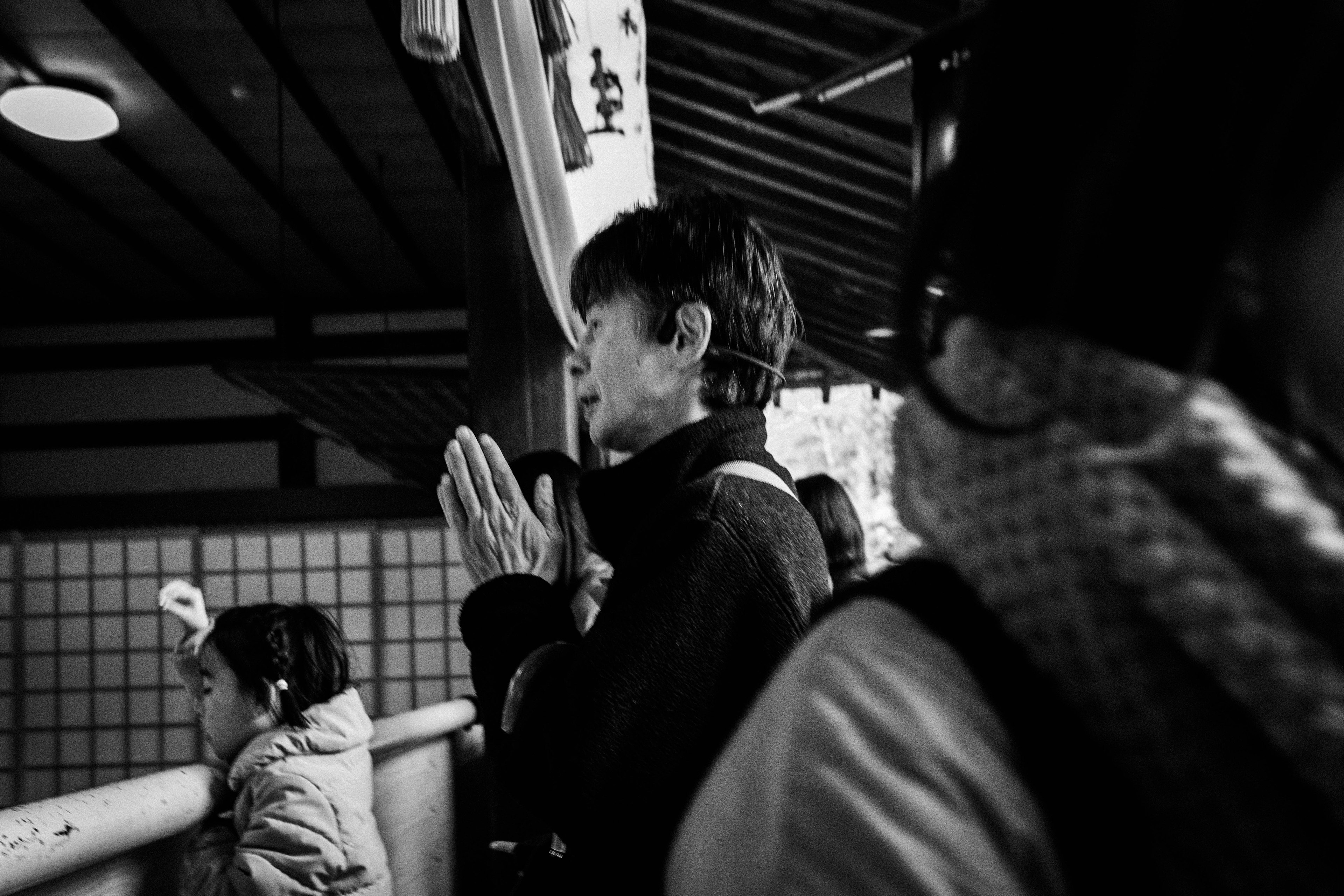 A black and white photo of a woman praying with hands together other people visible in the background