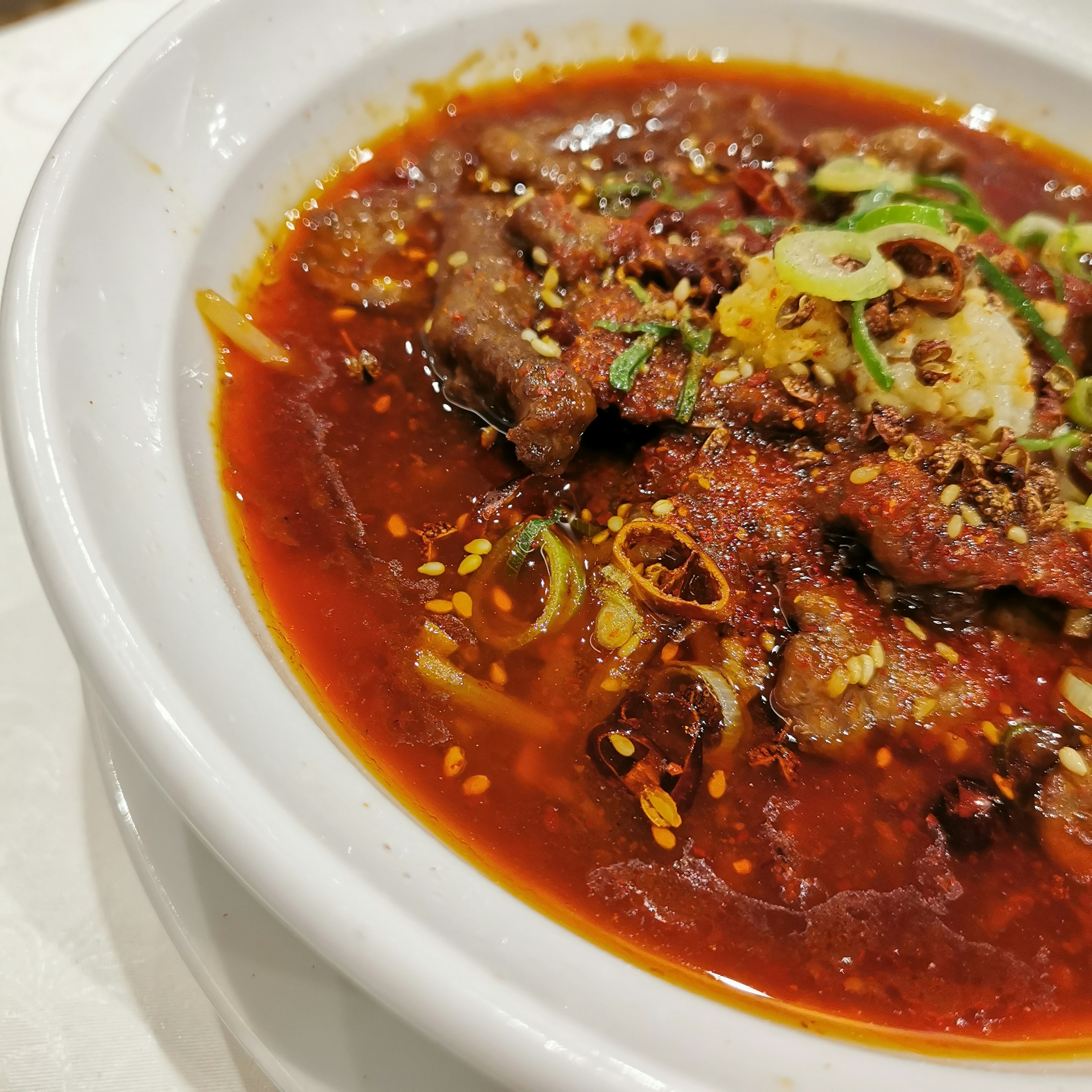 Un tazón de sopa roja picante con carne, decorado con cebollas verdes y condimento crujiente