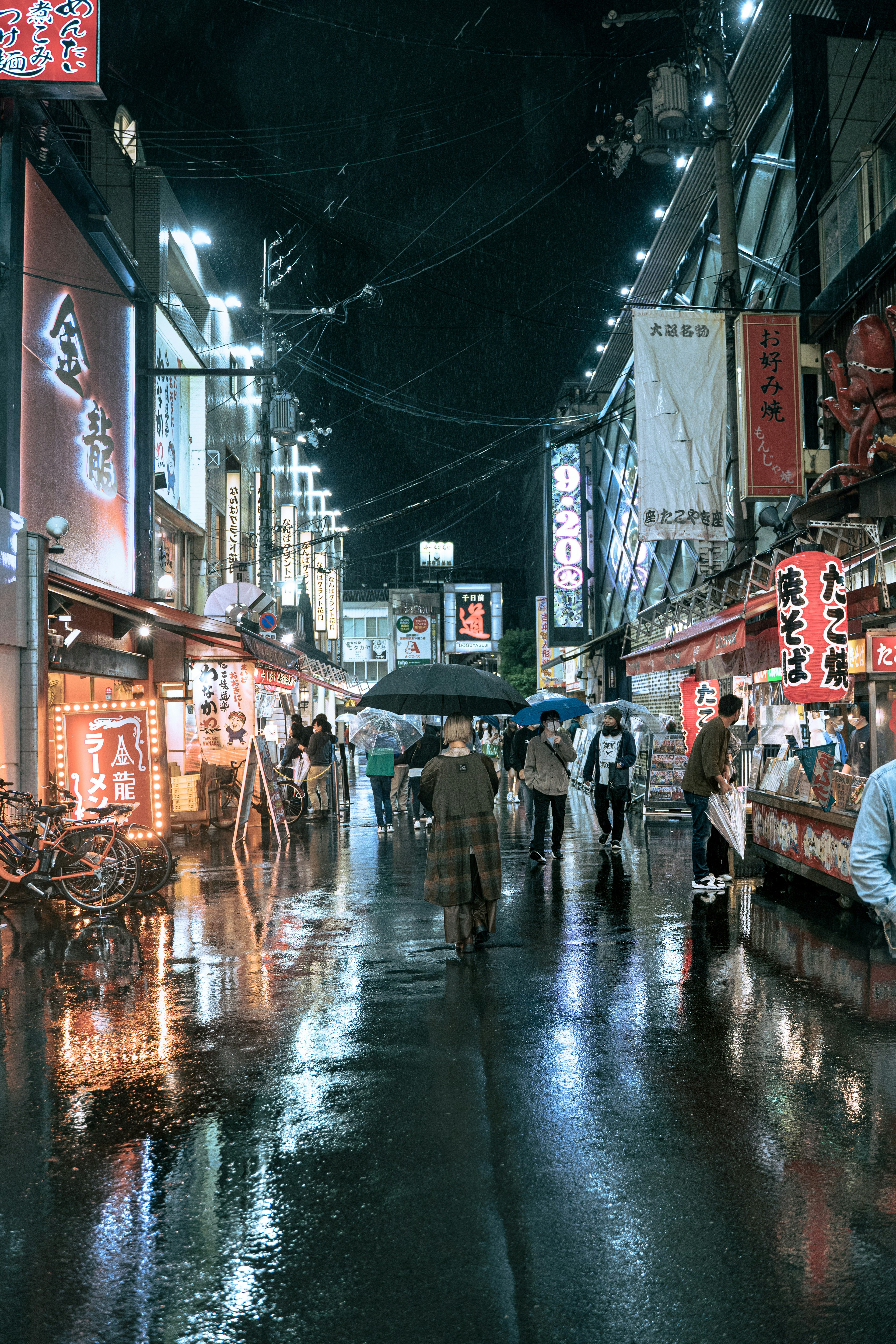 雨に濡れた夜の街並みに傘をさした人々が行き交う