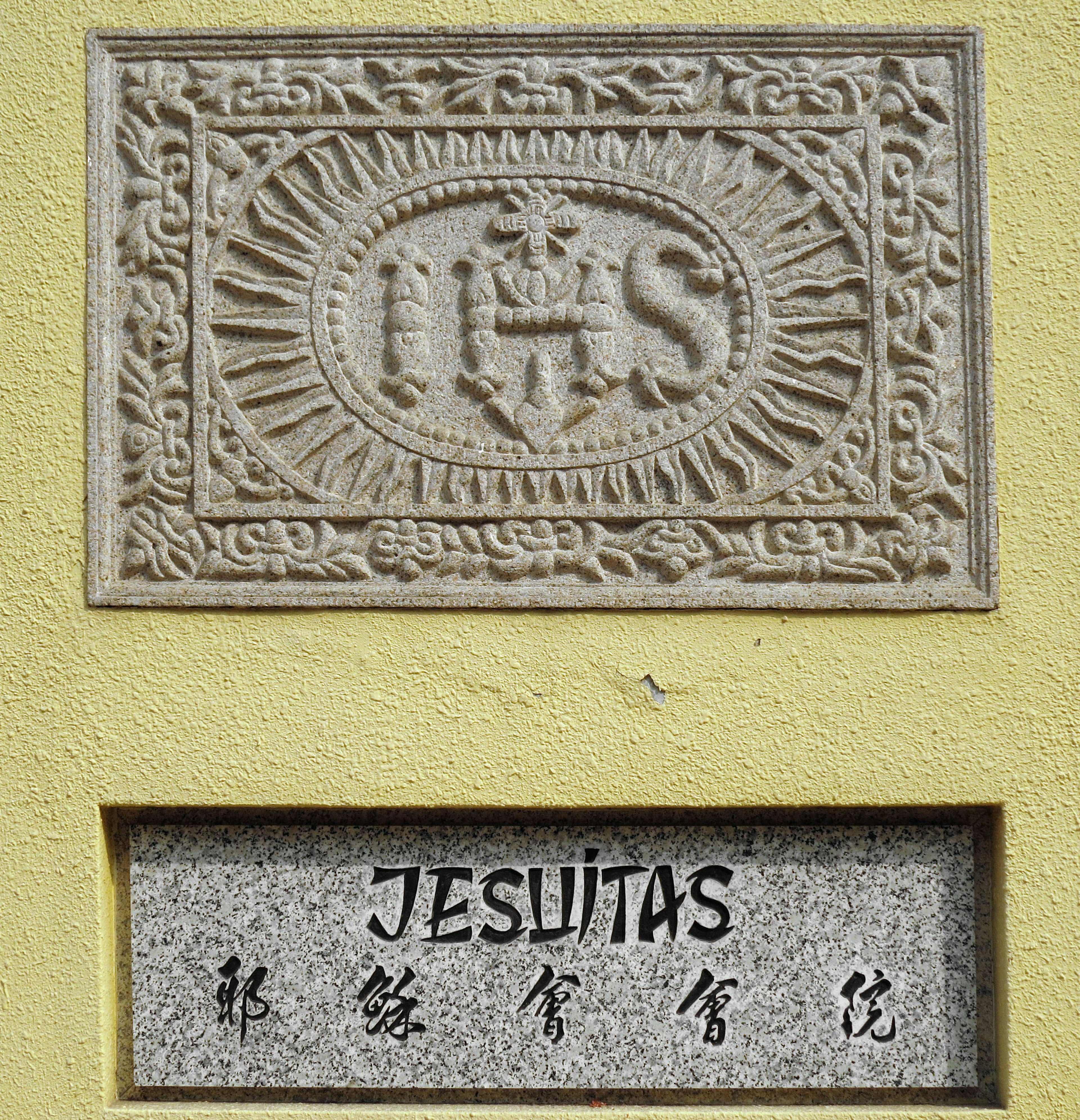 Eingravierte IHS-Emblem und Inschrift JESUITAS an einer gelben Wand