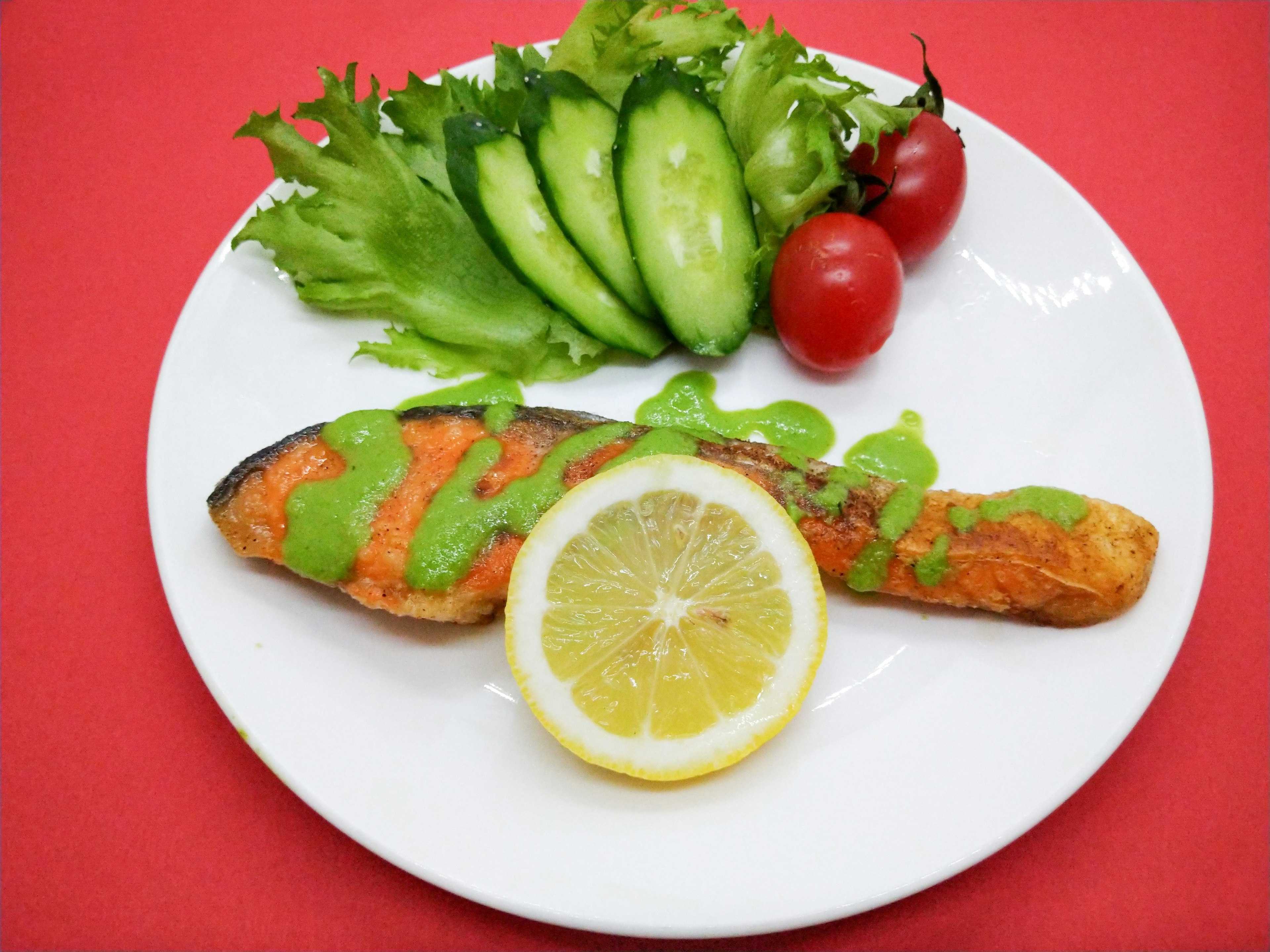 Ikan salmon panggang di piring putih dengan irisan lemon dan salad segar termasuk mentimun dan tomat ceri