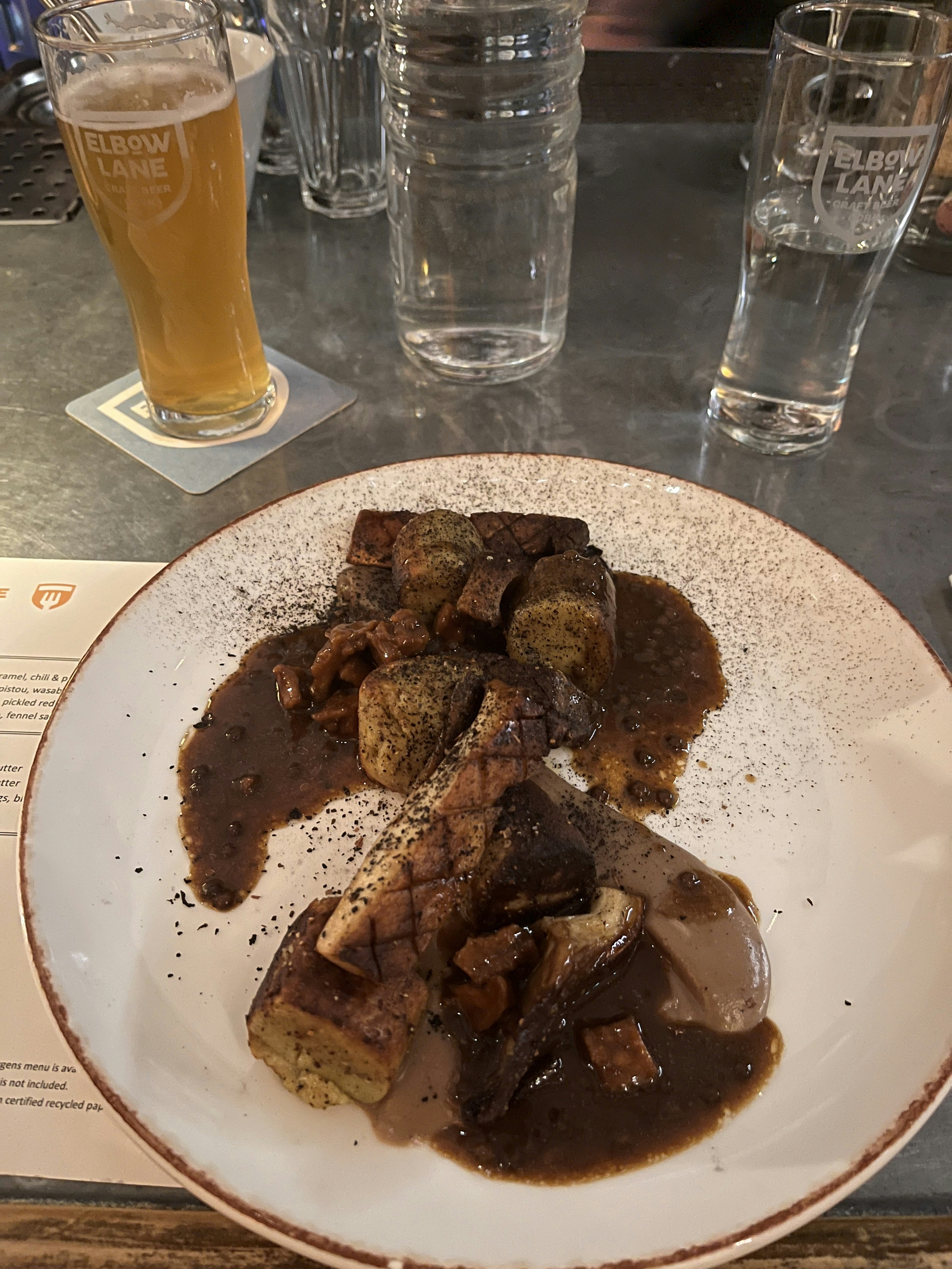 A plate of meat dish with sauce and garnishes alongside a glass of beer and water glasses in the background