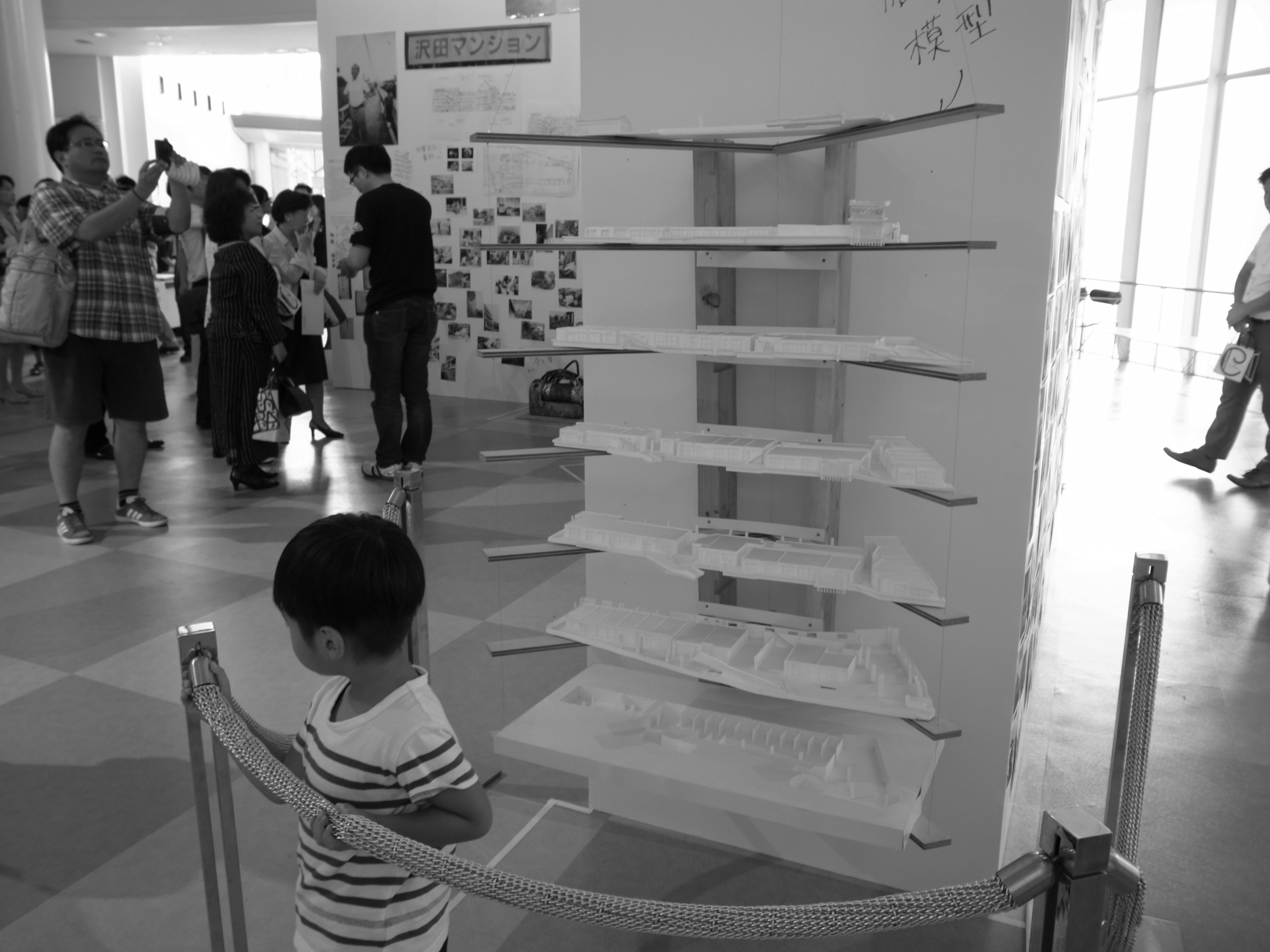 A child observing a white shelf structure in a black and white exhibition space with people in the background