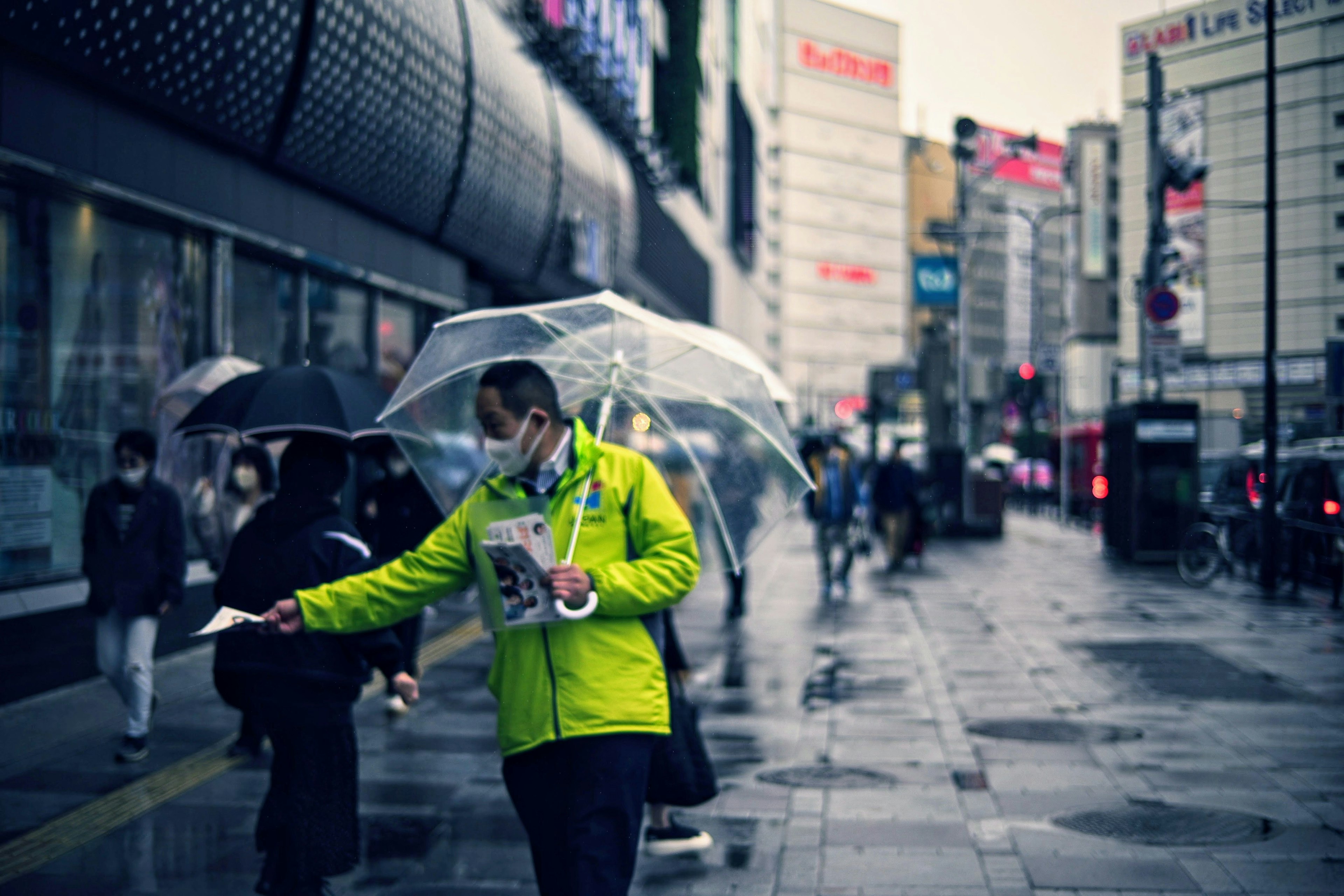 雨の中、透明な傘を持つ人が街を歩き、緑のジャケットを着ている