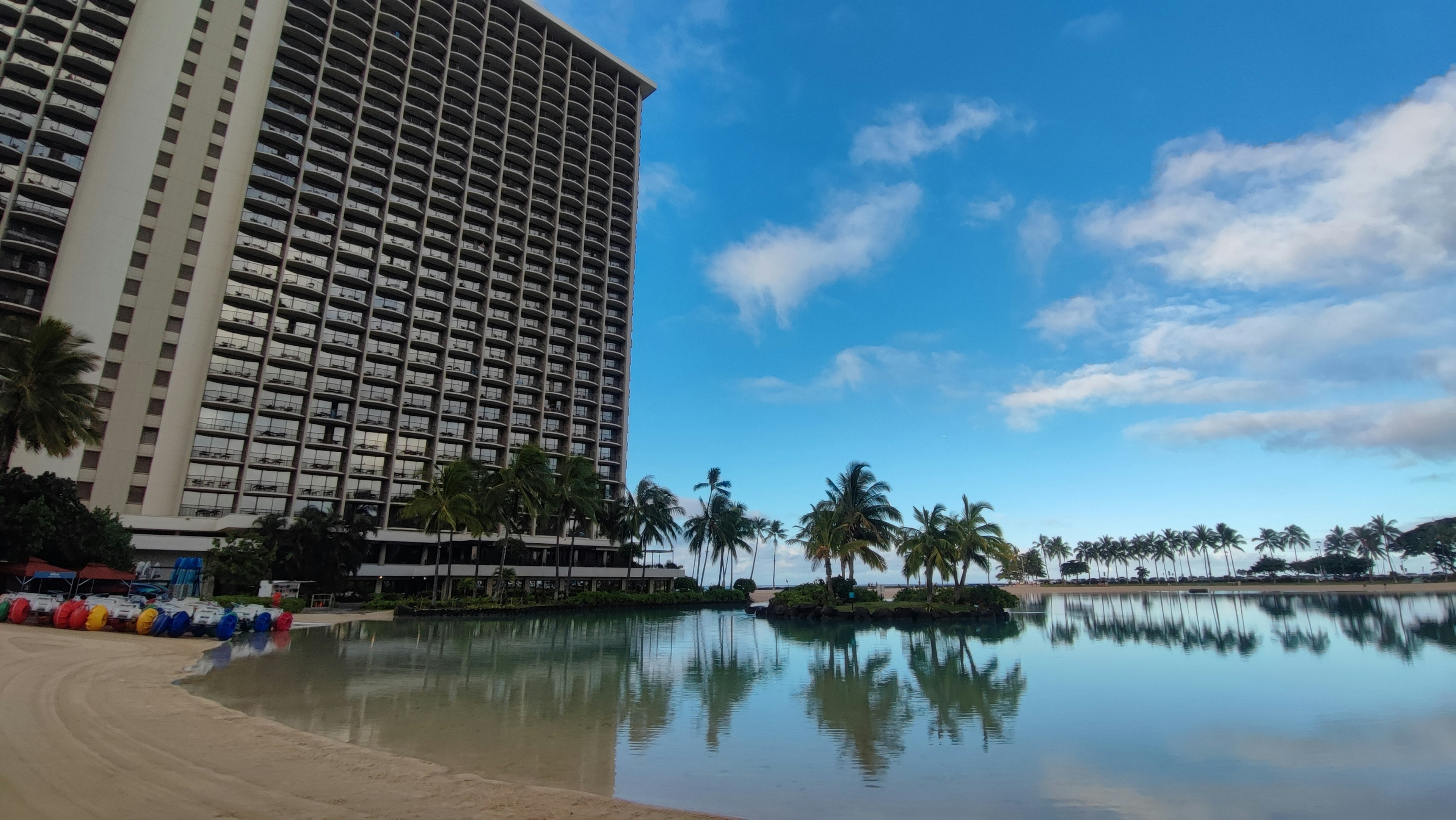 Vue panoramique d'un immeuble élevé sur une plage avec un ciel bleu clair et des palmiers reflétés dans l'eau calme