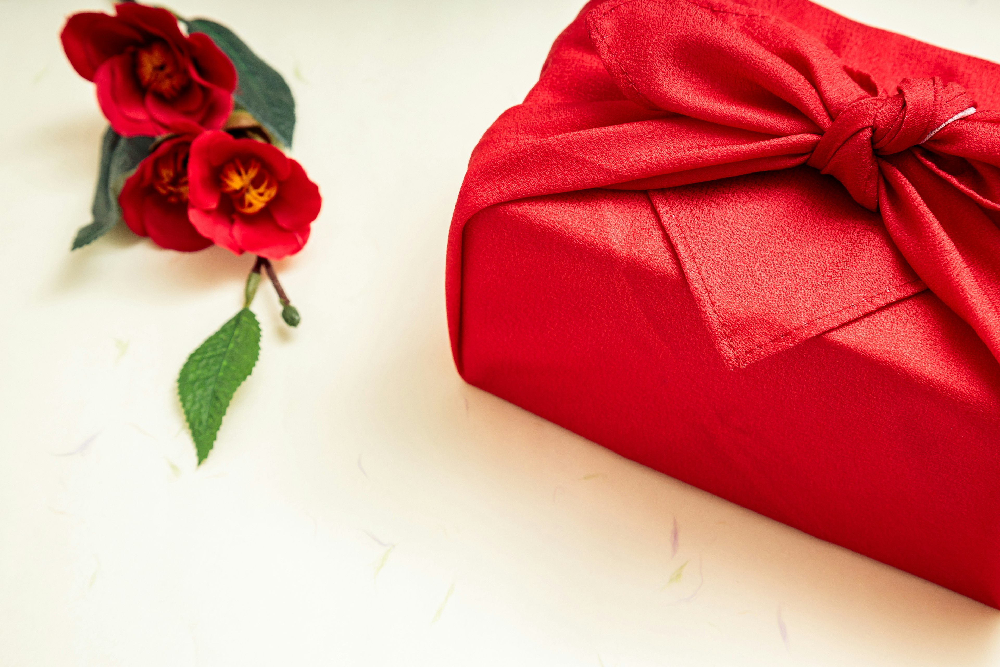 A red fabric-wrapped gift with a bow next to red flowers on a plain background