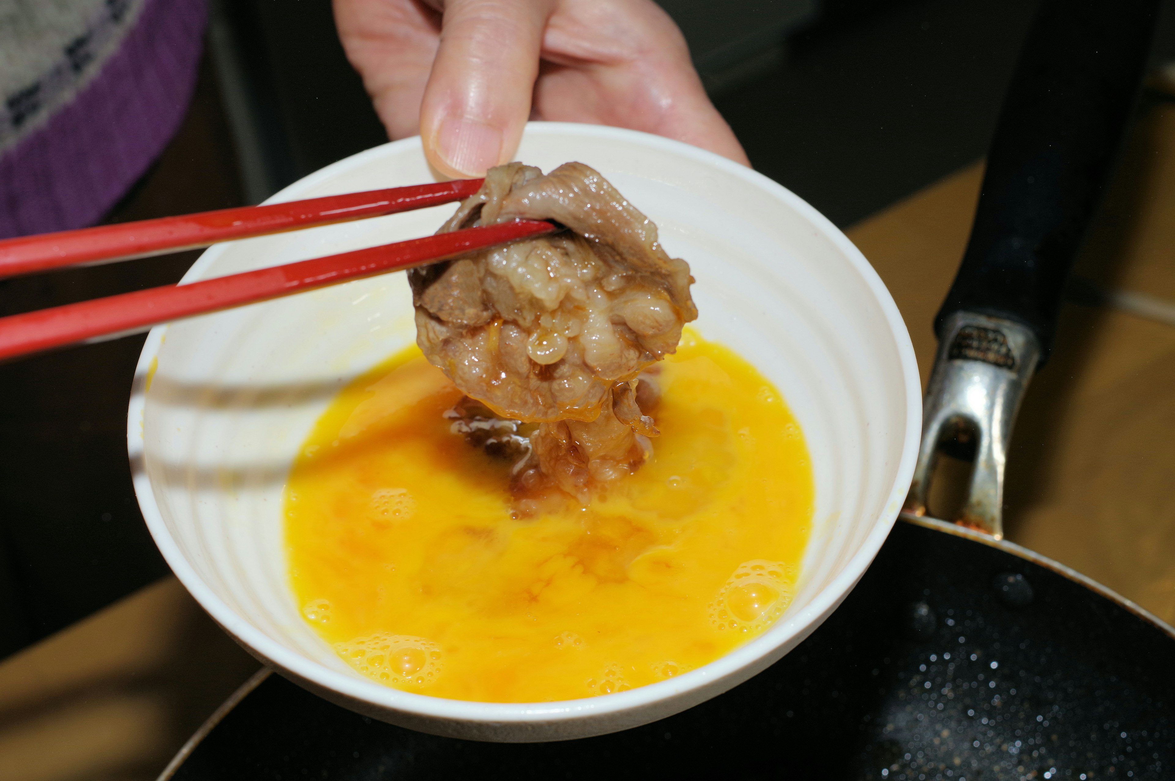 A hand using chopsticks to dip meat into a bowl of egg mixture