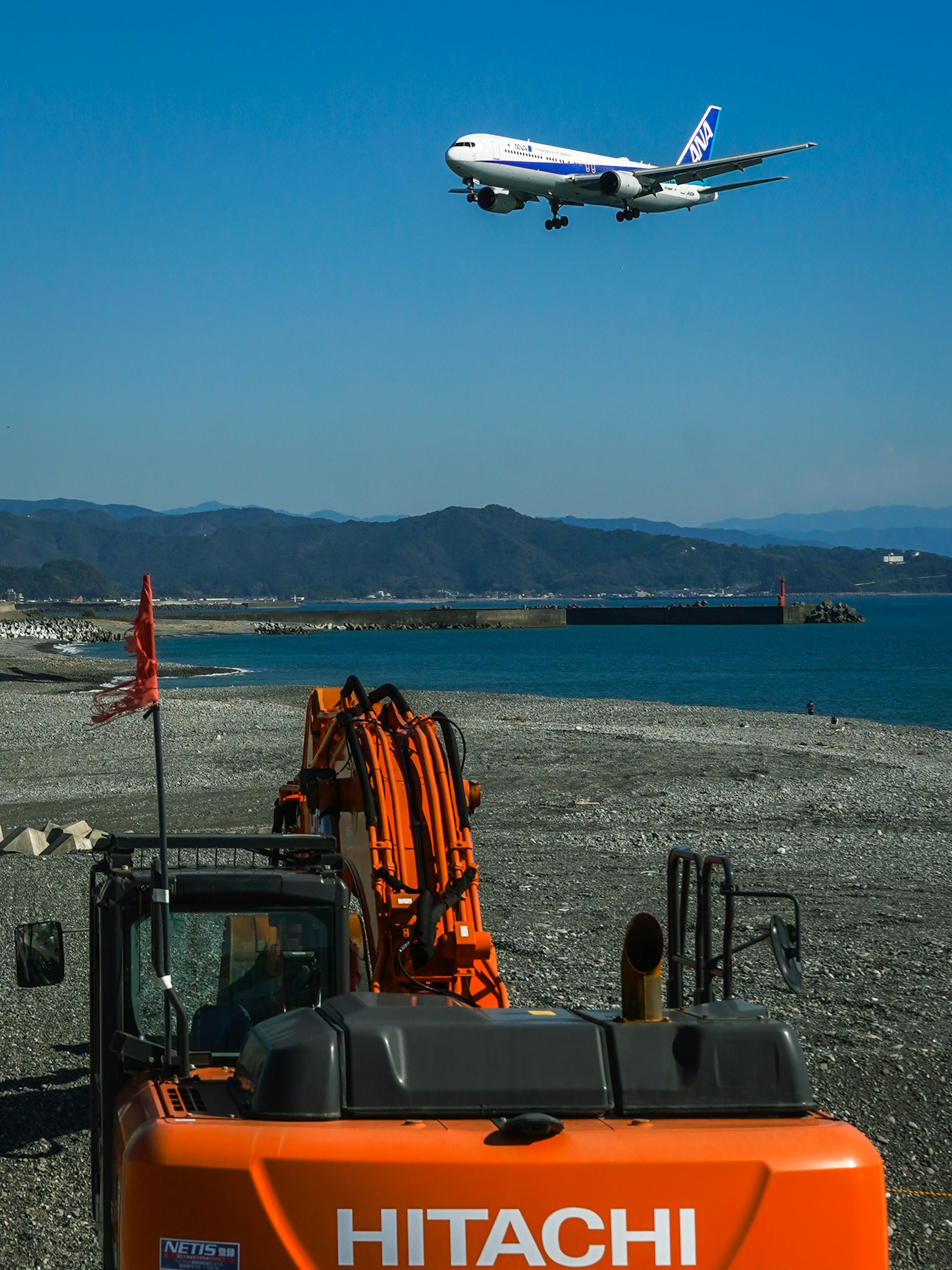 海辺で飛行機が飛ぶ中 HITACHIの重機が目立つ風景