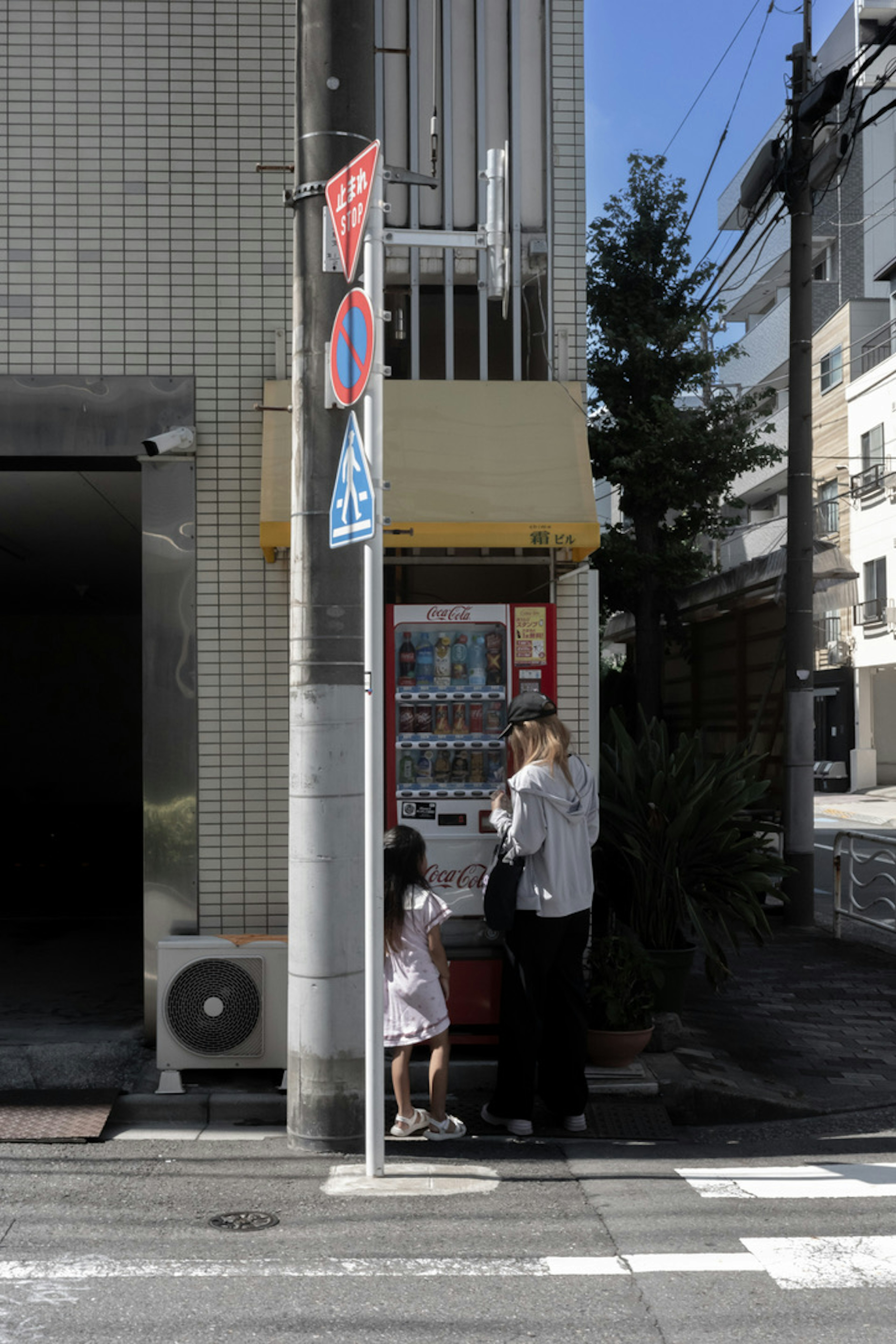 Silhouette eines Elternteils und eines Kindes an einem Automaten an einer Straßenecke mit umliegenden Gebäuden