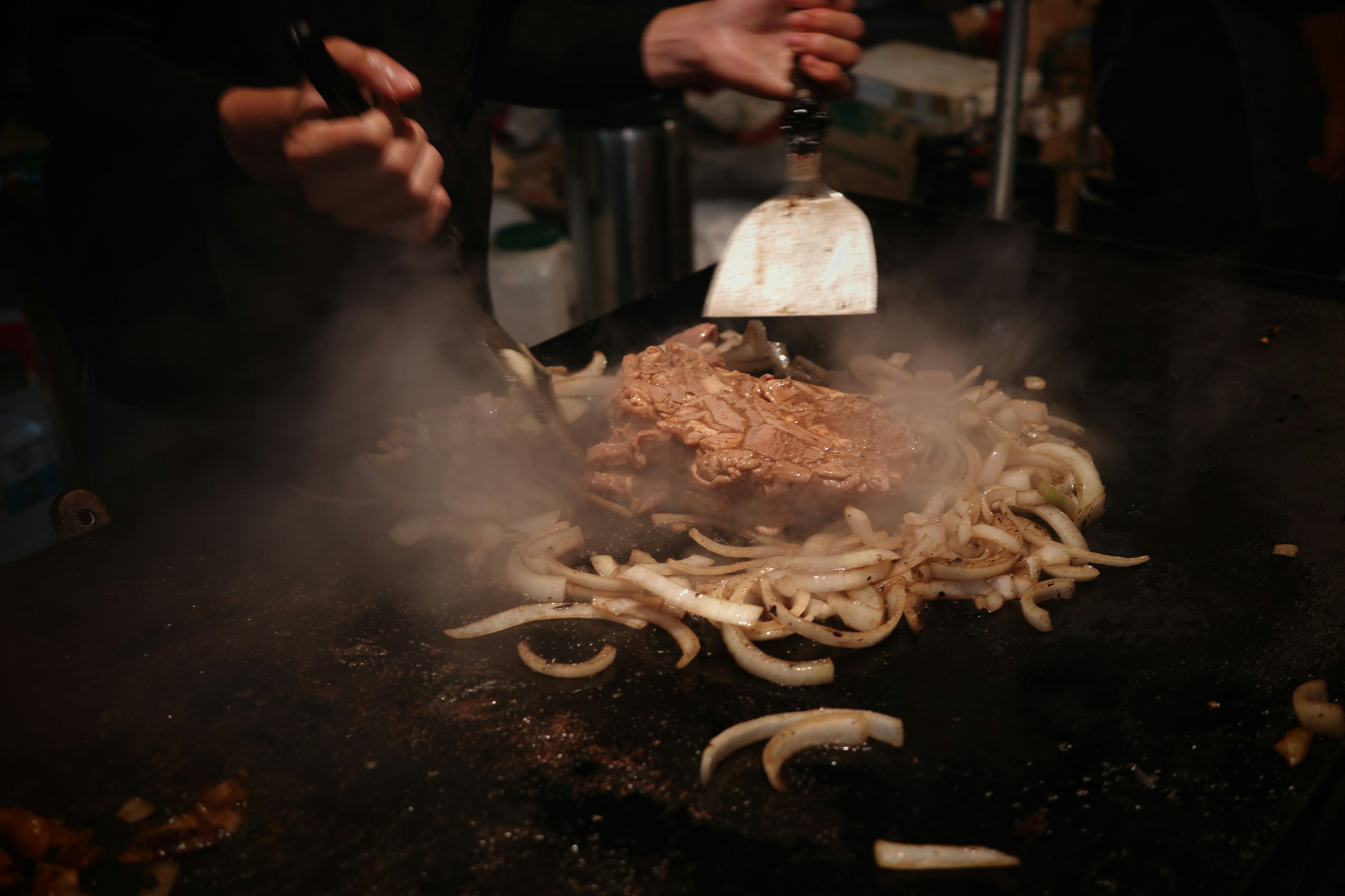 煙が立ち上る鉄板の上で肉と玉ねぎを調理している手元