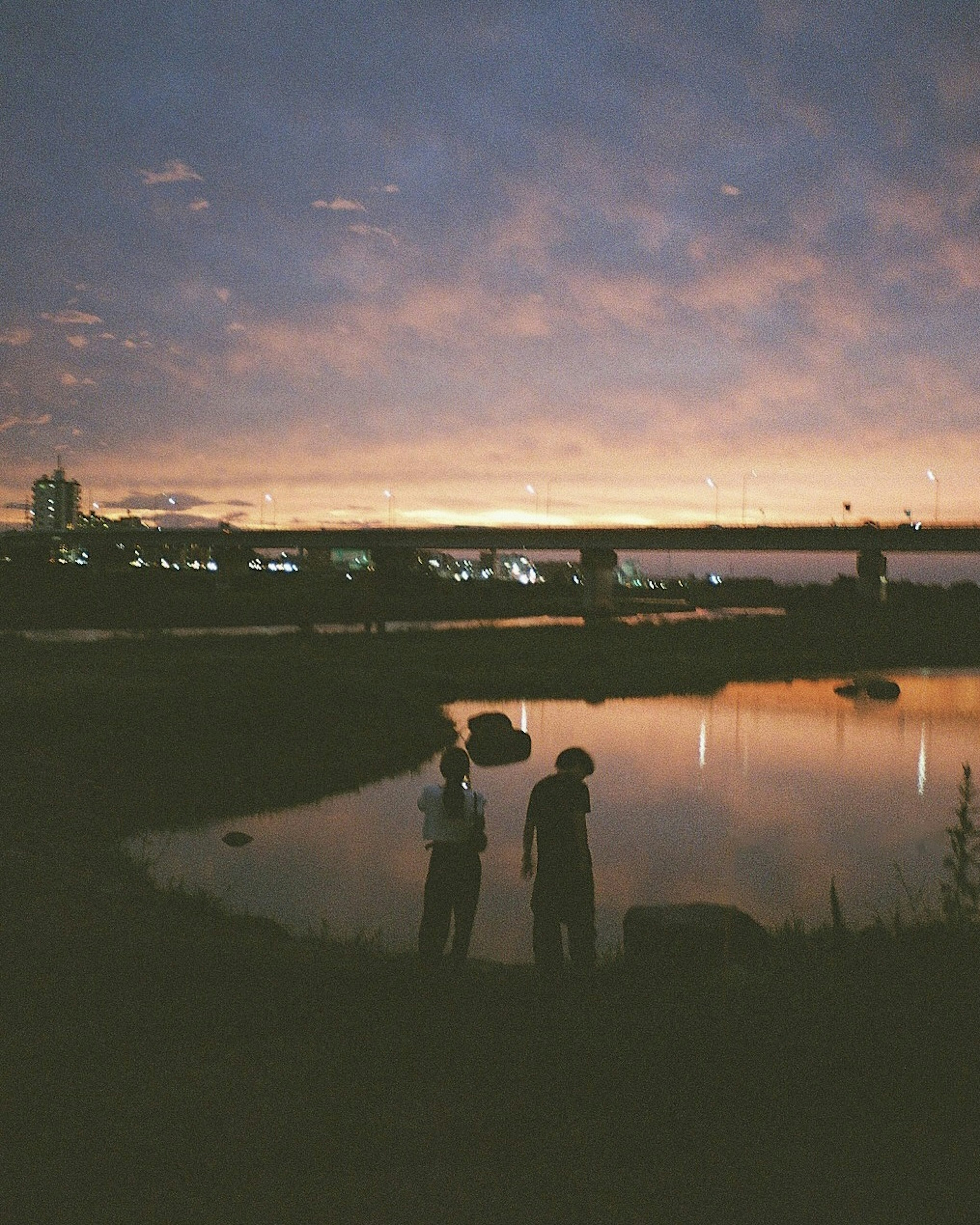 Two people standing by a riverside at sunset with reflections on the water