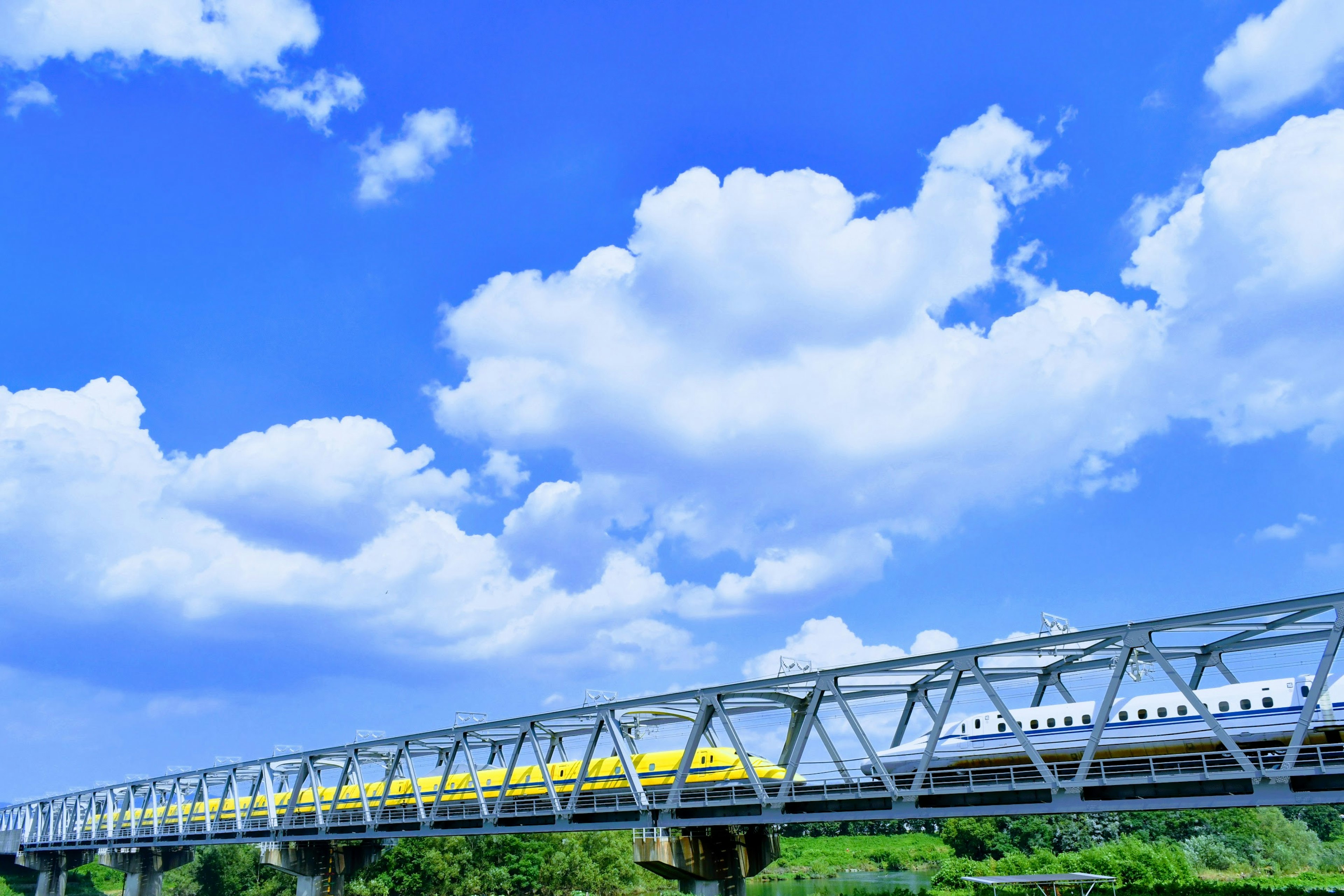 A yellow train crossing a steel bridge under a blue sky with fluffy white clouds