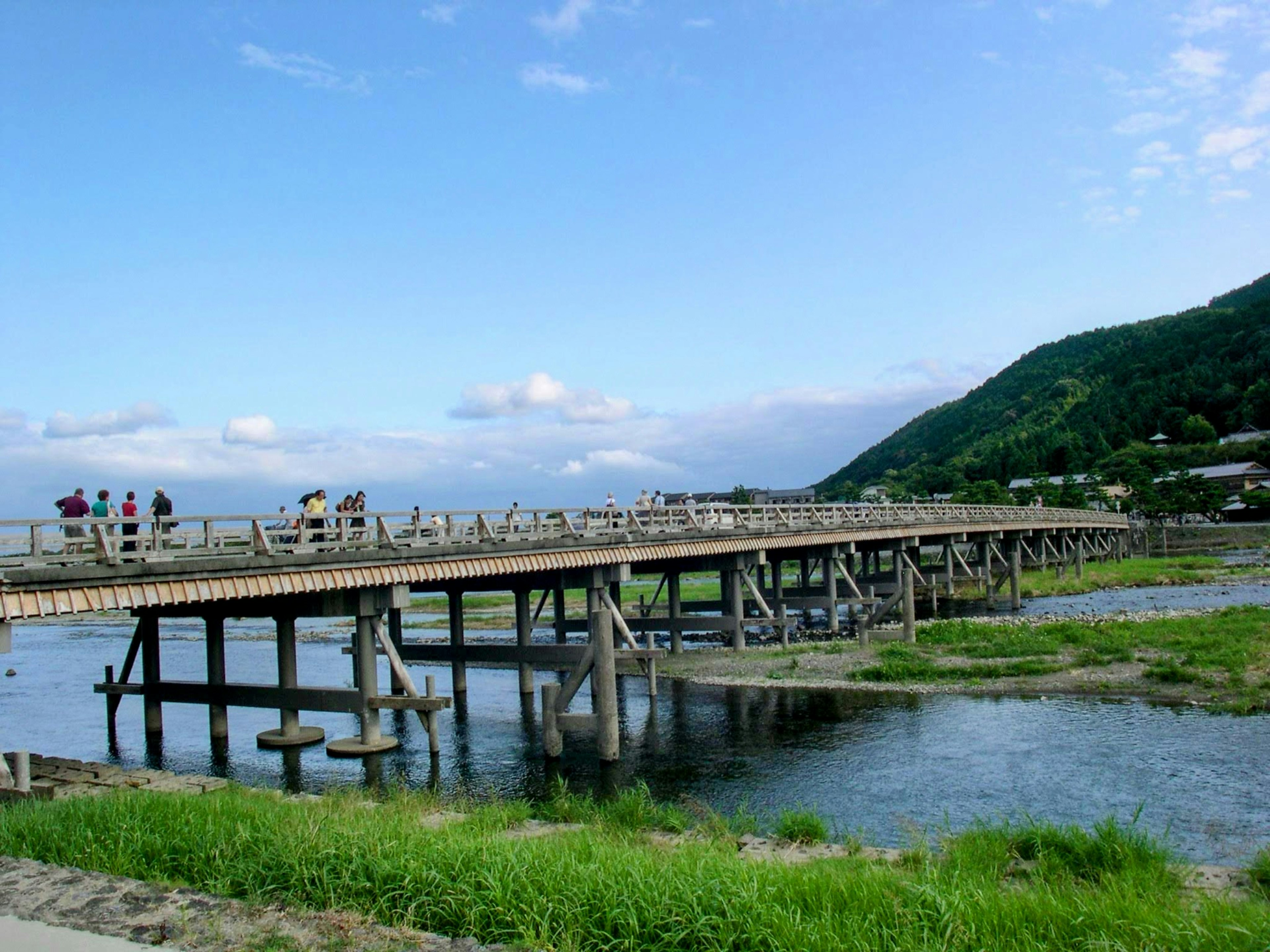 Jembatan kayu yang melintasi sungai dengan rumput hijau dan langit biru