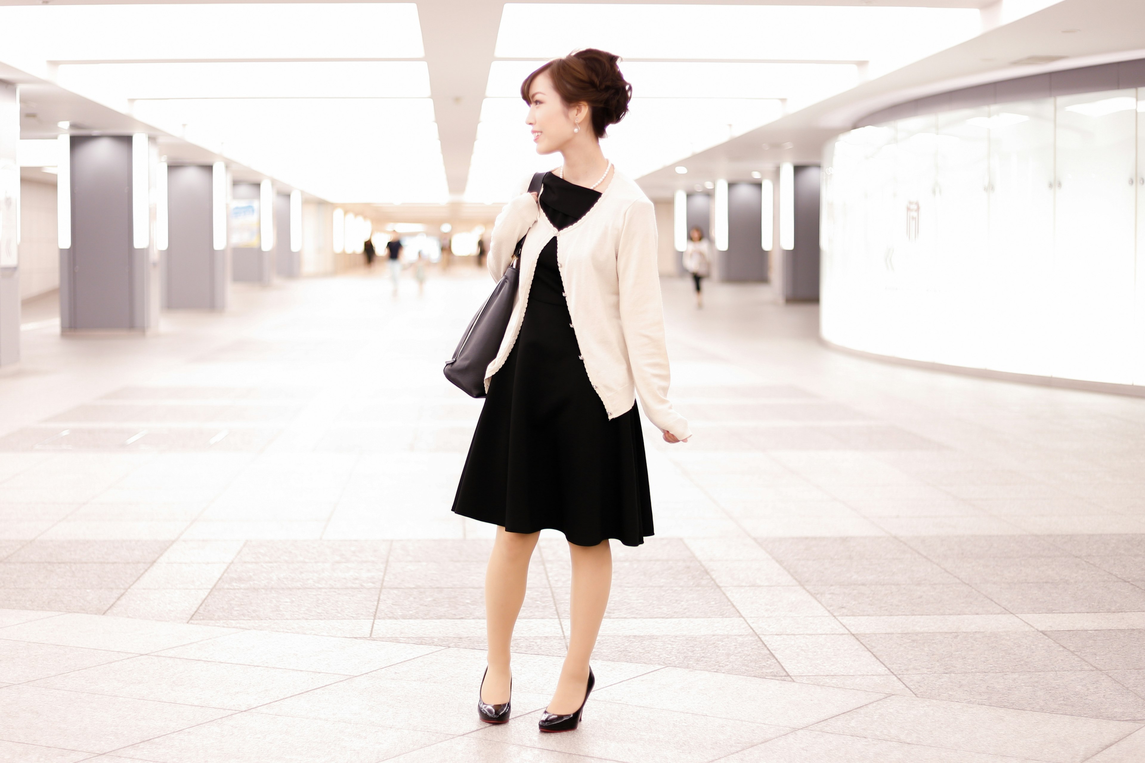 Femme en robe noire et veste blanche posant dans un espace moderne