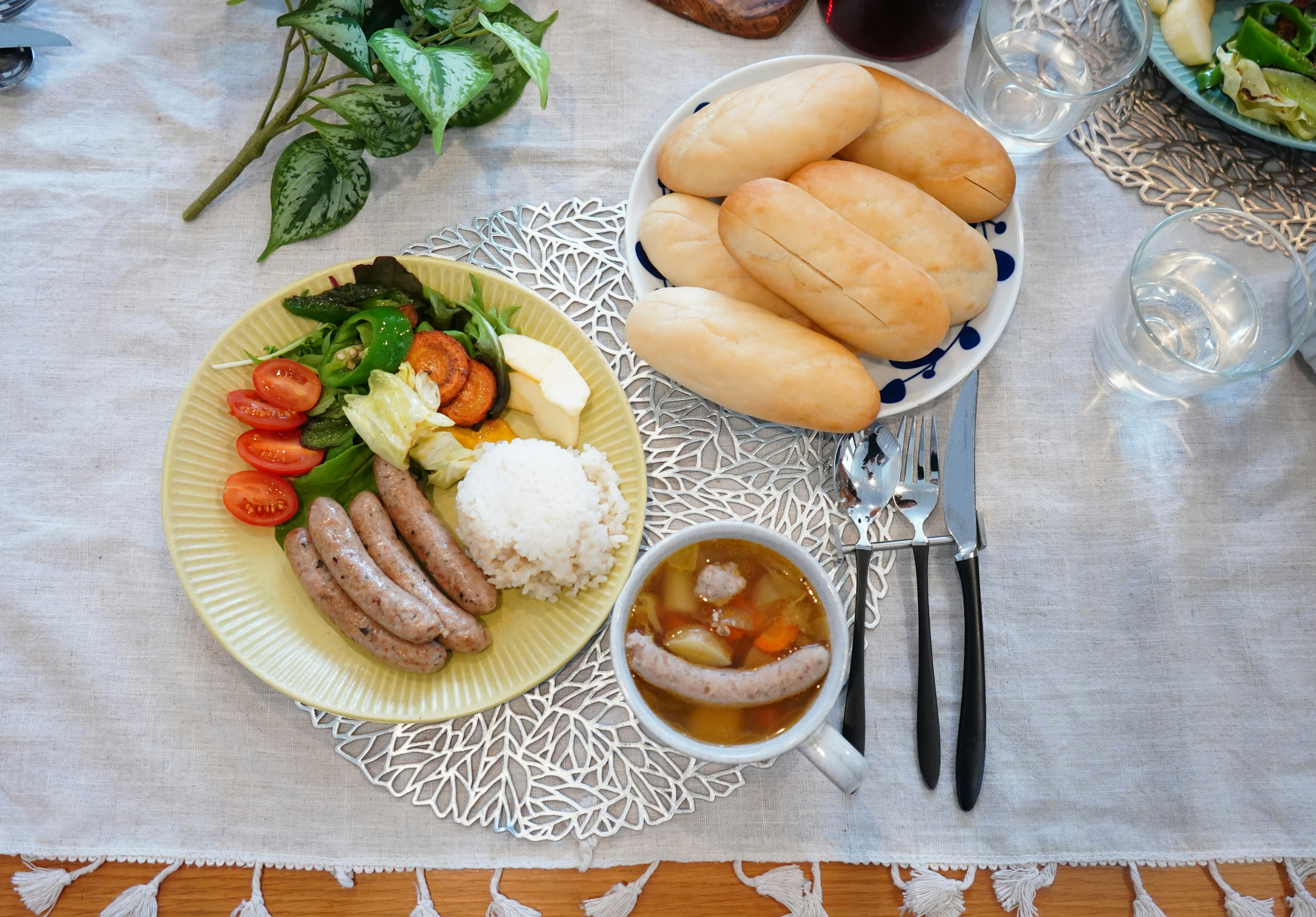 A plate of sausages, vegetables, and rice with a bowl of soup and a plate of bread on a dining table