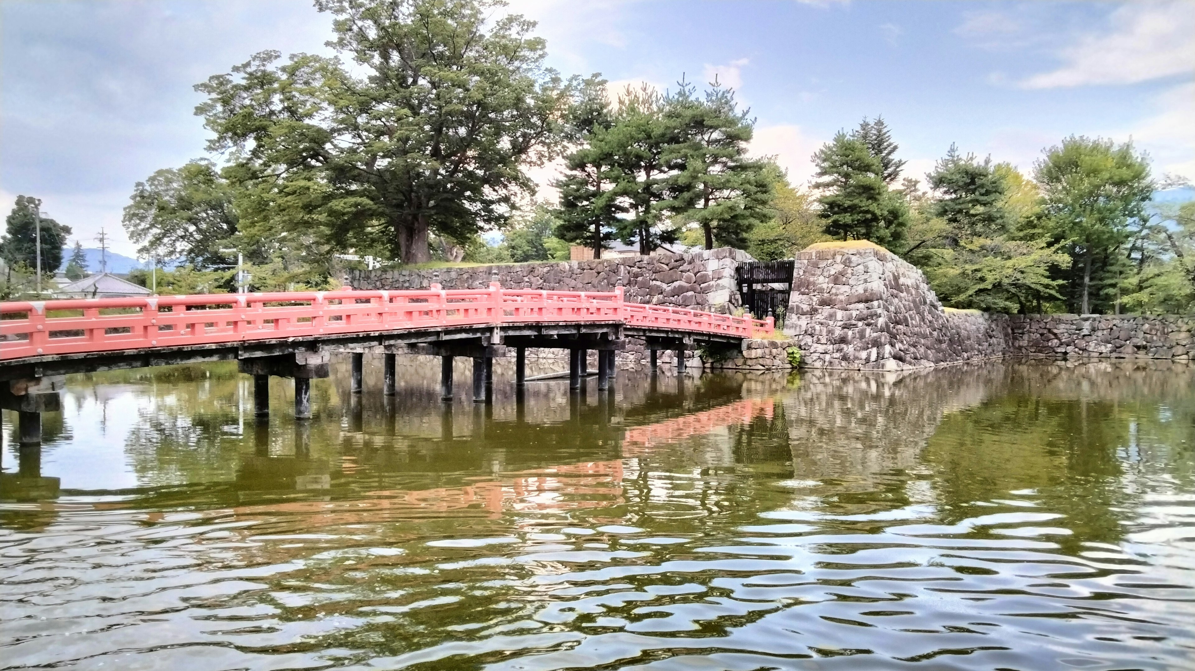 Vista escénica de un puente rojo y un terraplén de piedra reflejado en agua tranquila