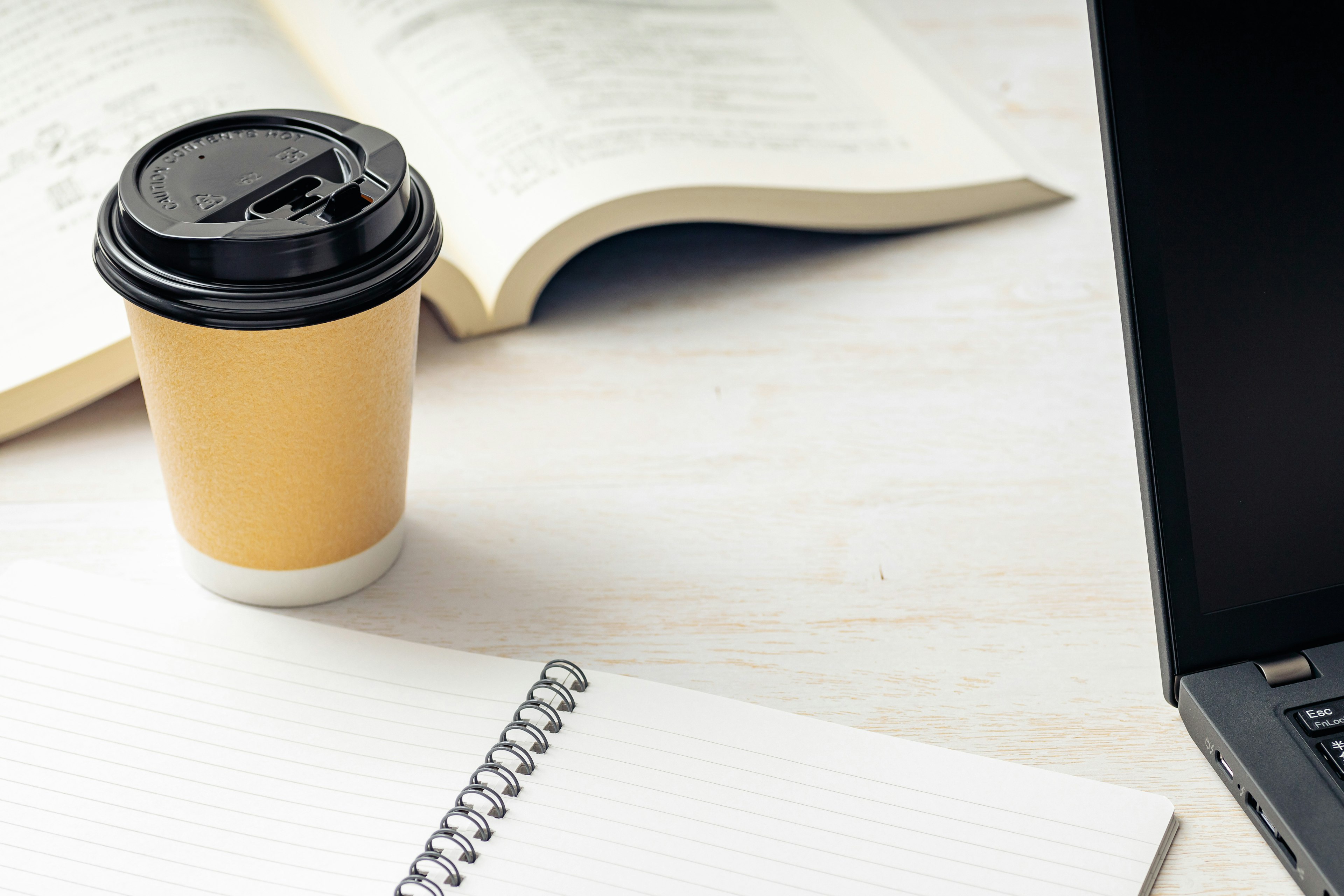 Kaffeetasse und Laptop auf einem Schreibtisch mit offenen Büchern und einem Notizbuch in einer einfachen Arbeitsumgebung