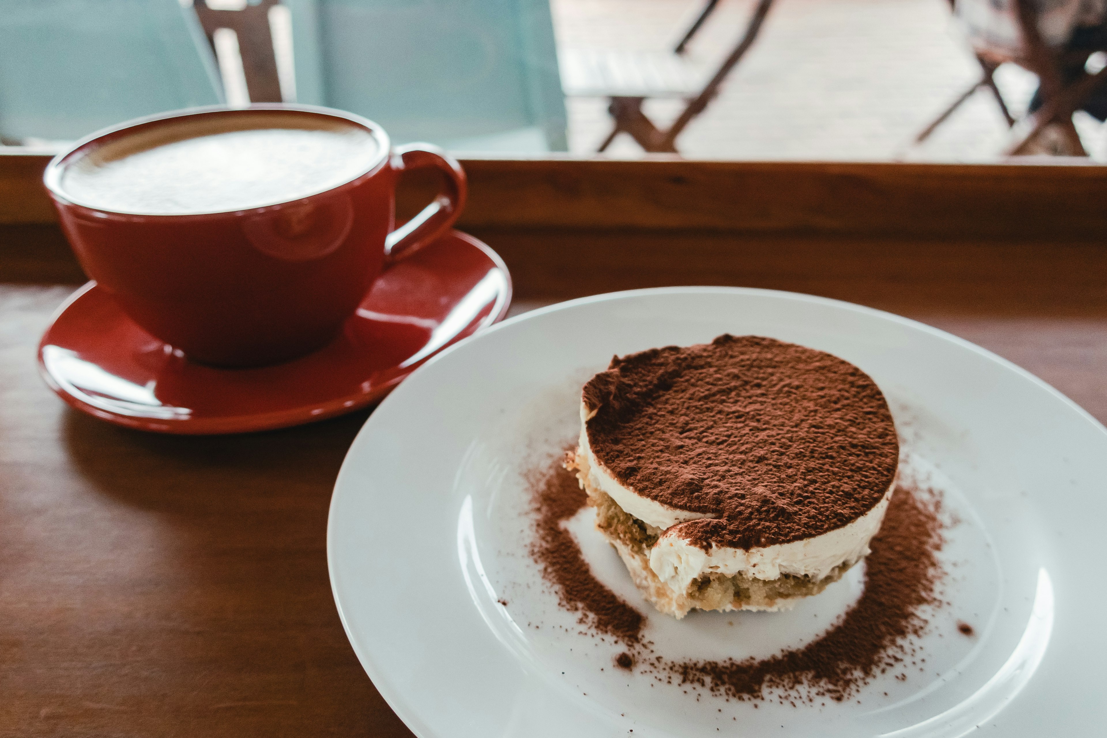 A red coffee cup beside a plate of tiramisu on a wooden tray in a cafe setting