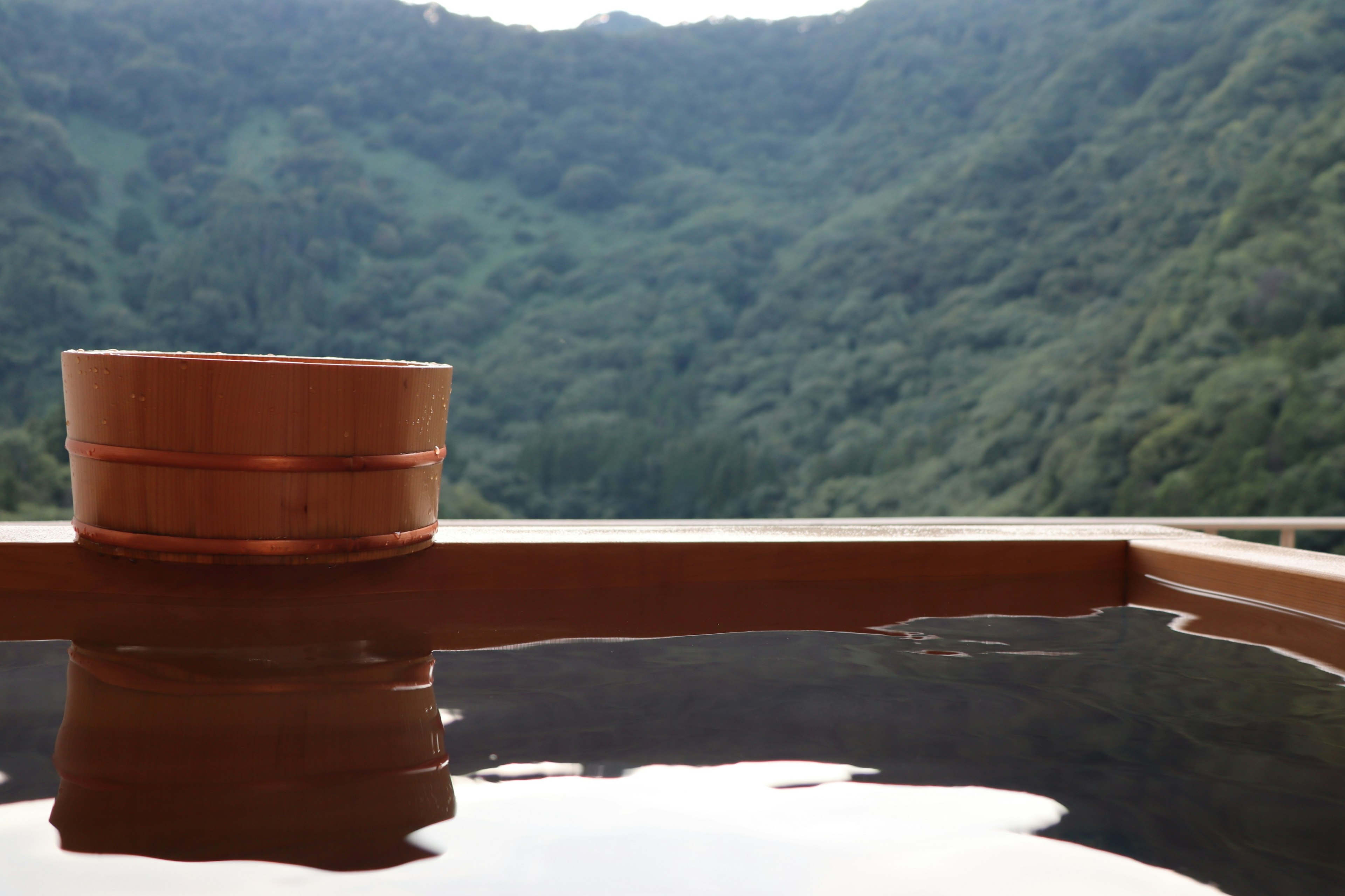 Wooden hot spring tub with serene water surface and green mountains in the background