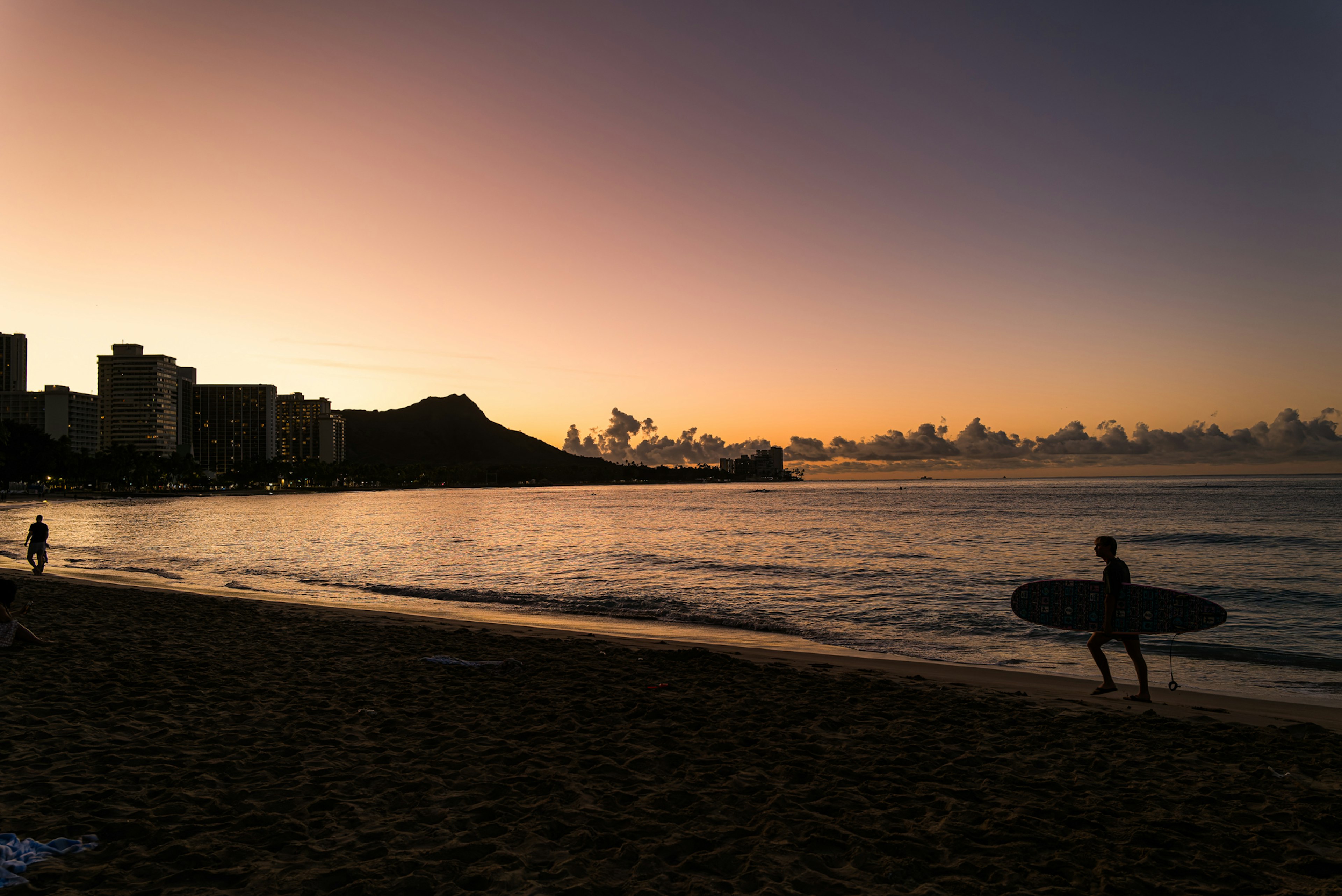 夕焼けのビーチでサーフボードを持ったサーファーが歩いているハワイの風景