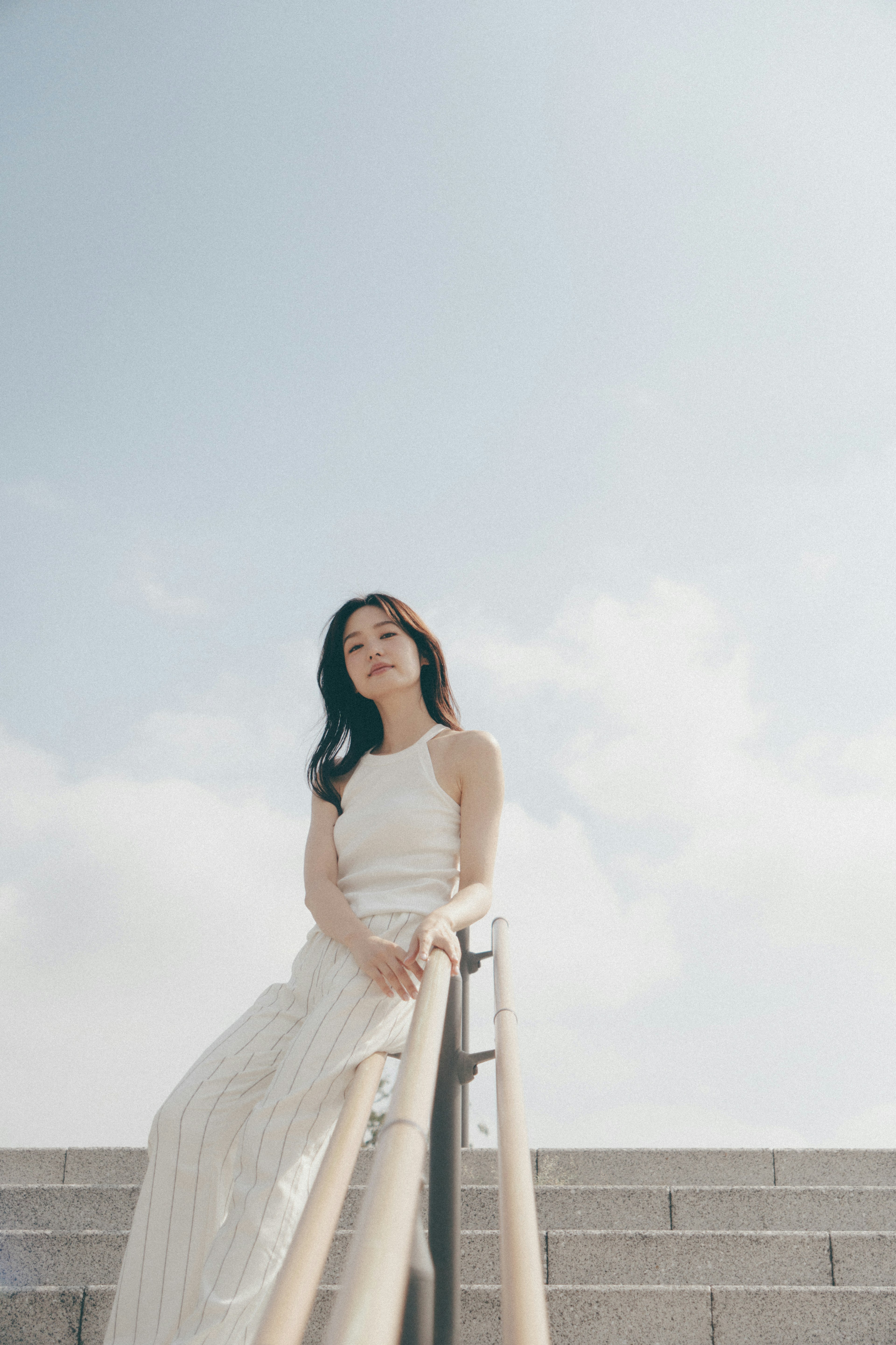 A woman in a white dress leaning against a handrail on stairs under a blue sky