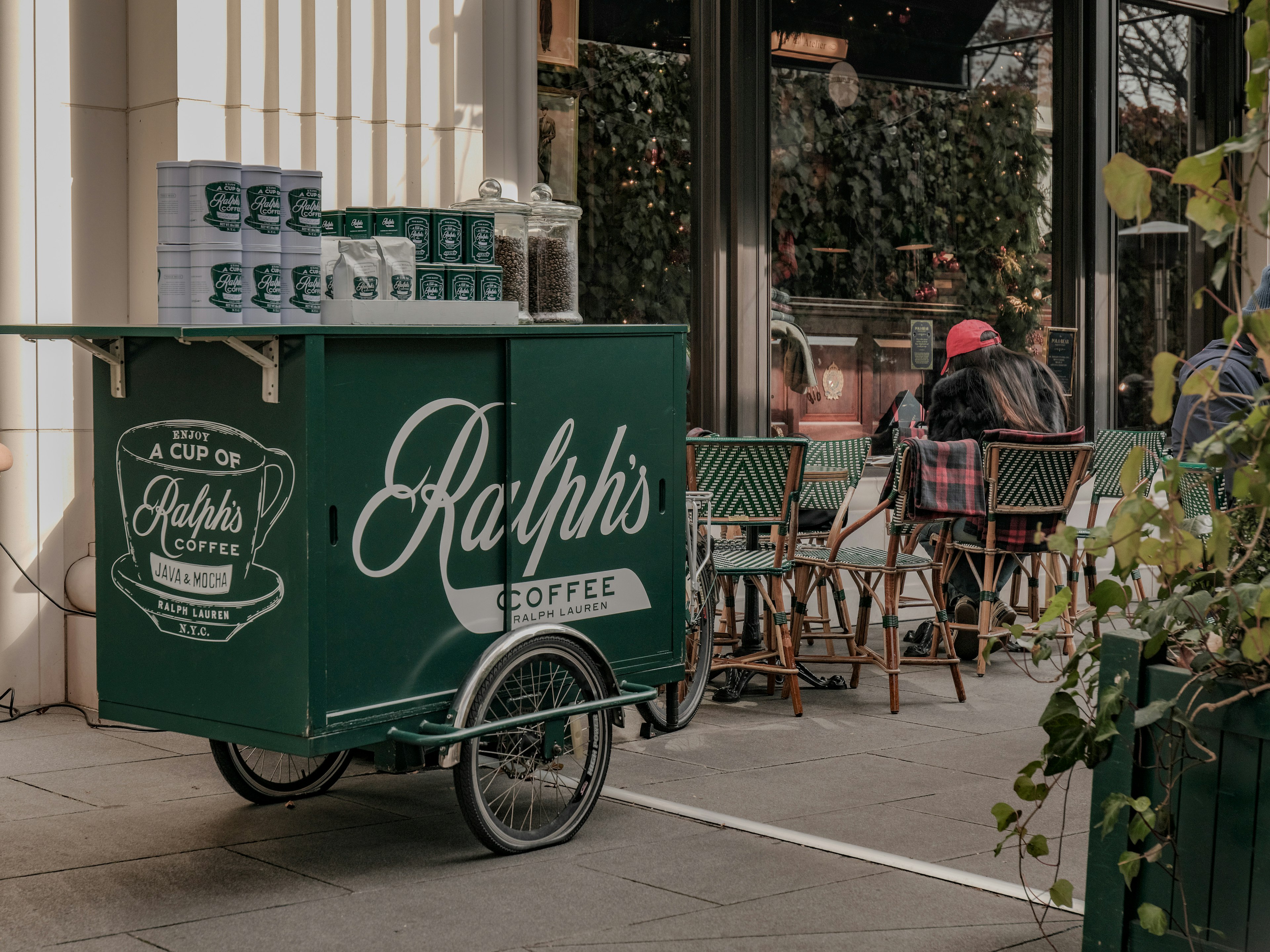 Green coffee cart with 'Ralph's Coffee' logo in front of outdoor café tables