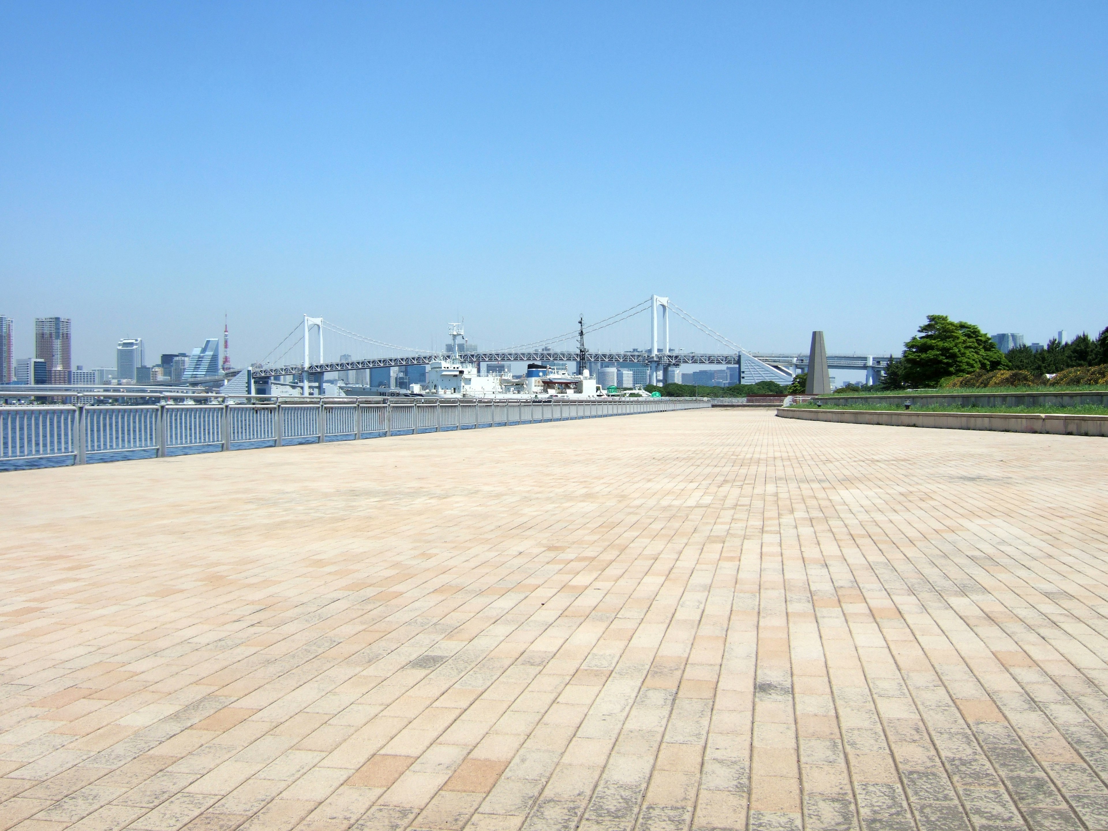 Plaza carrelée spacieuse sous un ciel bleu clair avec un pont et des bateaux au loin