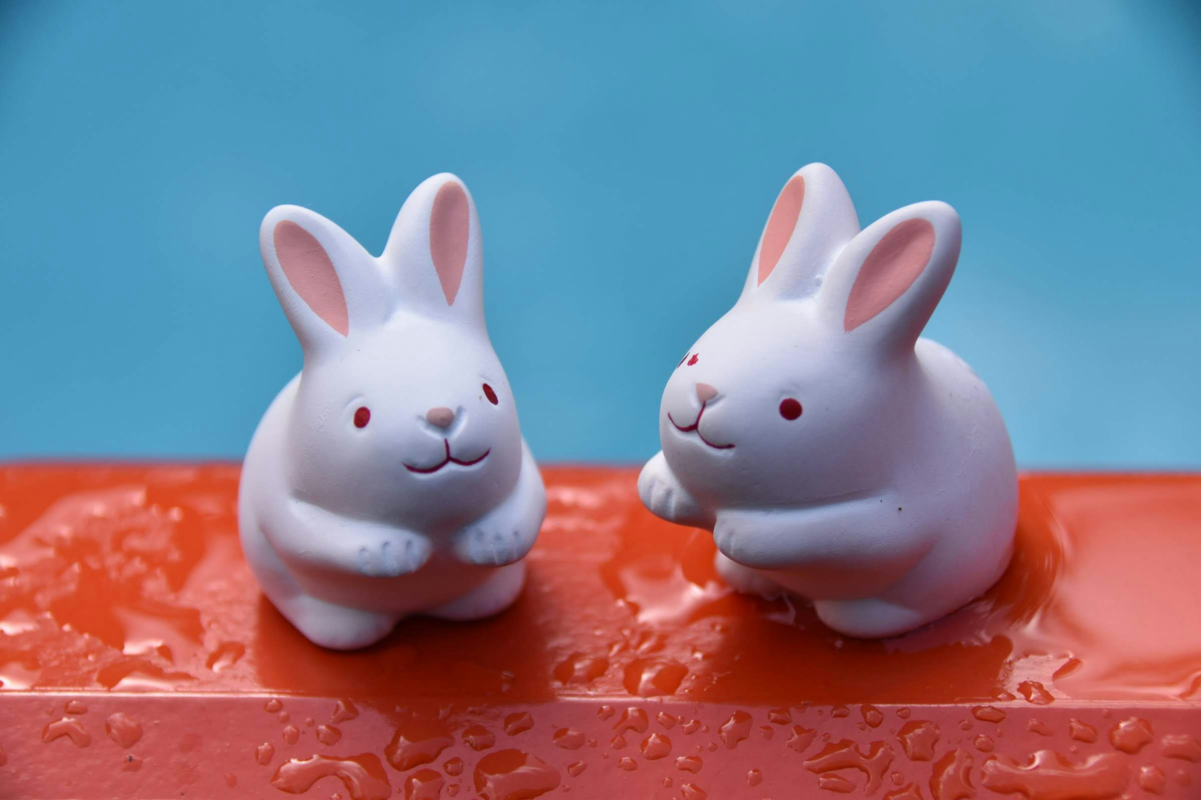 Dos figuras de conejos blancos sentadas sobre una superficie roja con un fondo azul