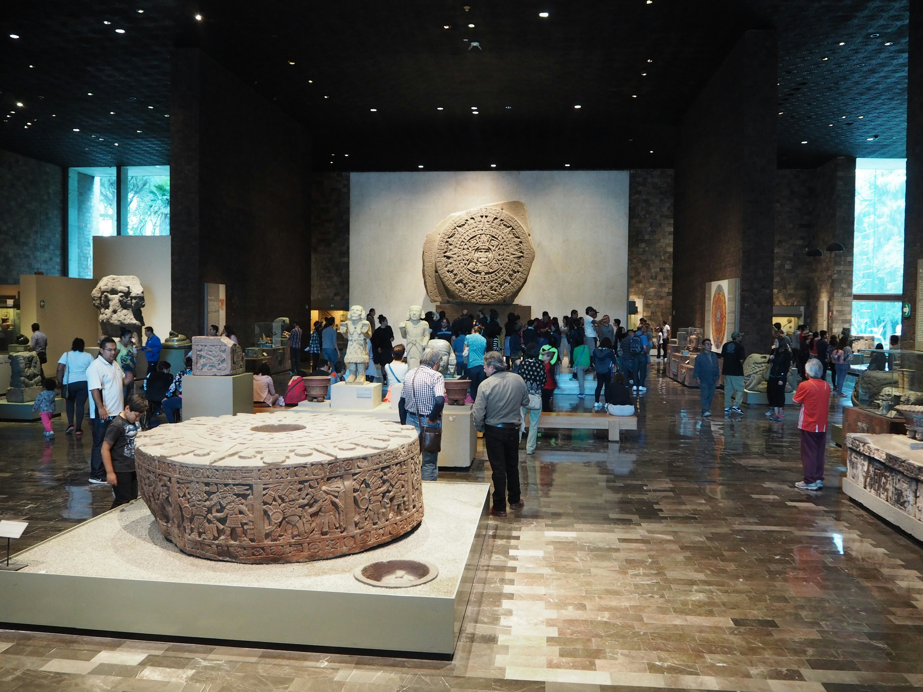 A spacious museum exhibit with a large circular sculpture in the center surrounded by various artifacts and many visitors exploring the area