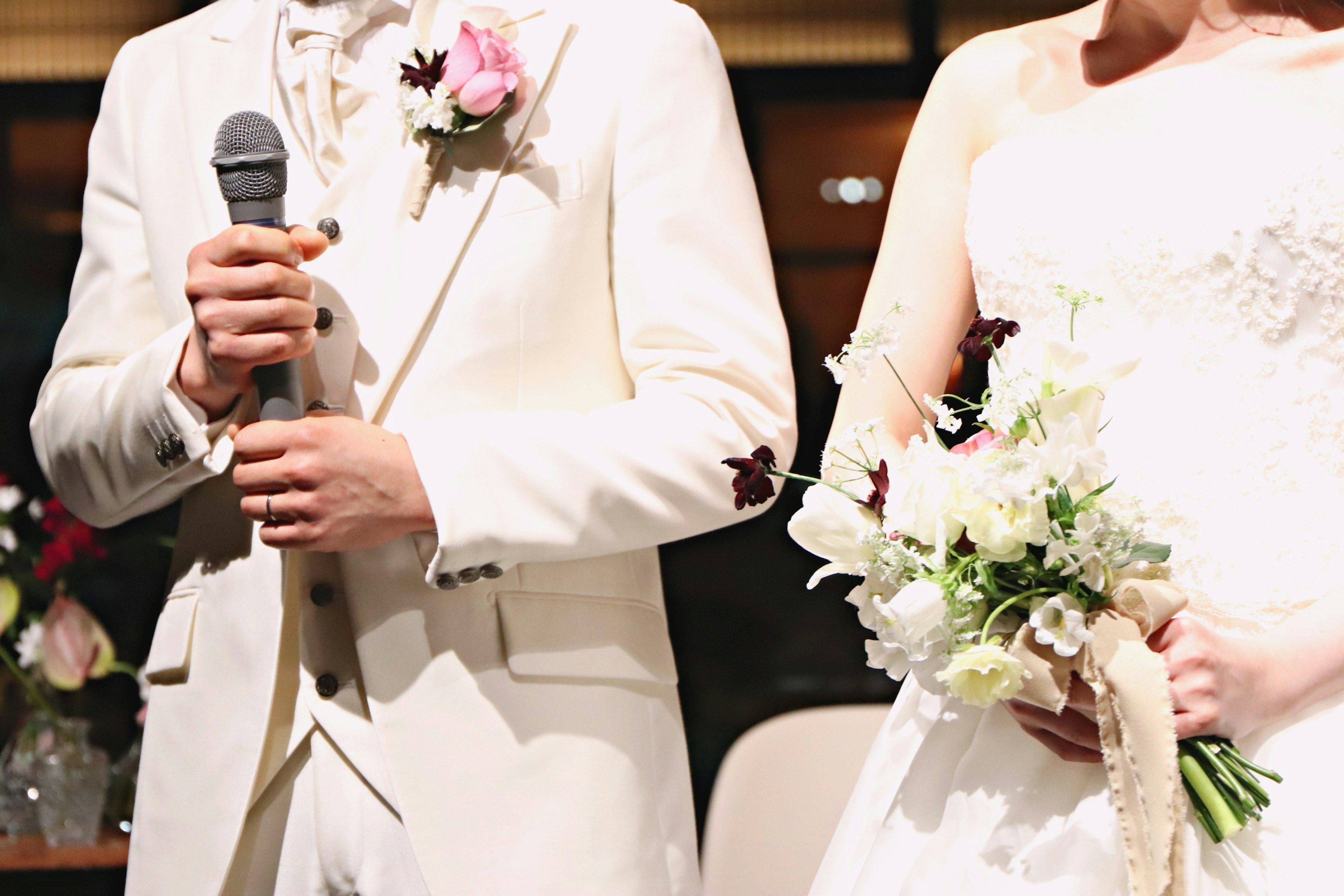 Man in white tuxedo holding a microphone and woman in wedding dress holding a bouquet