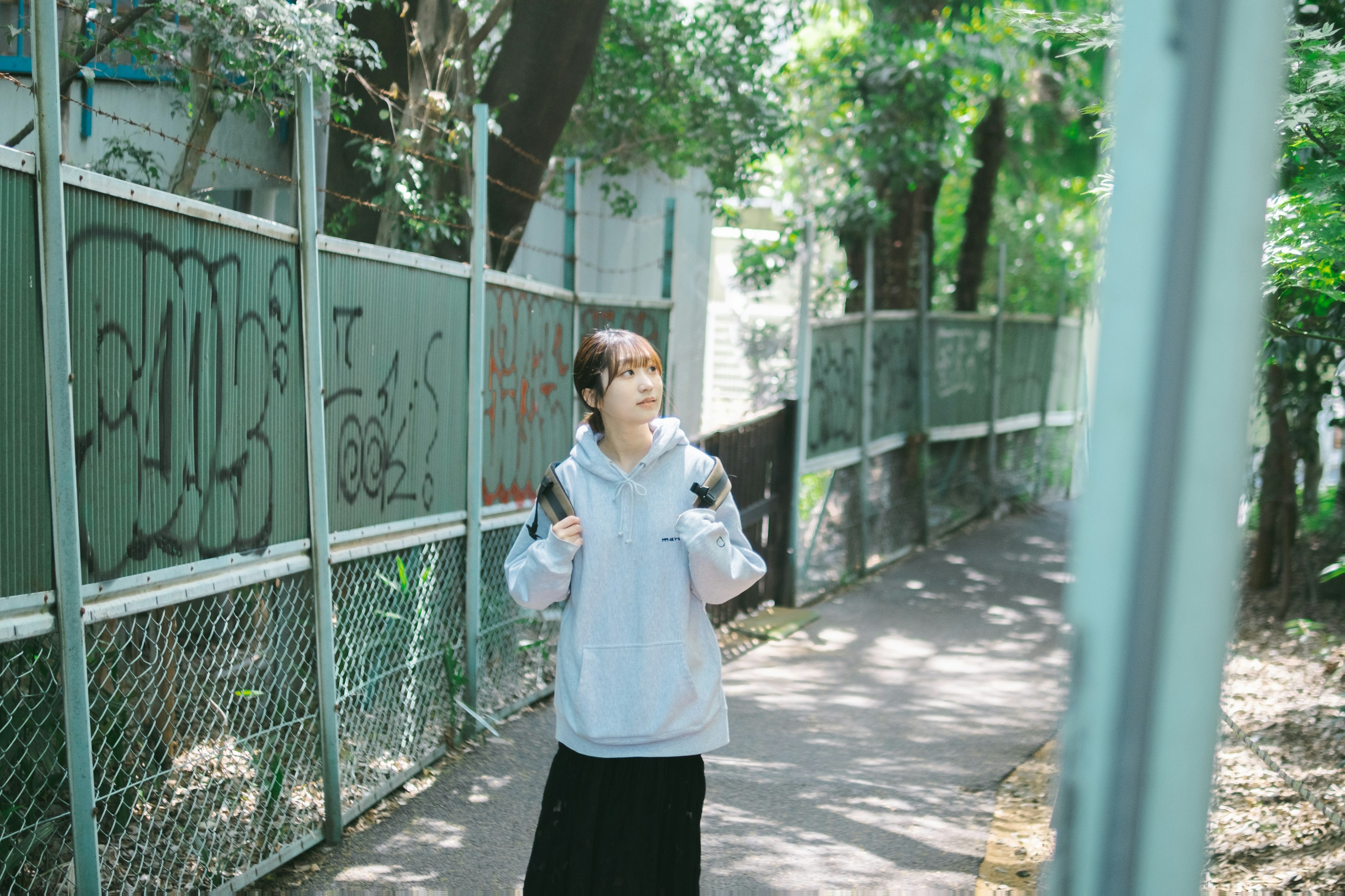 Woman walking on a park path wearing a gray hoodie and black skirt surrounded by green trees and a fence sunlight filtering through