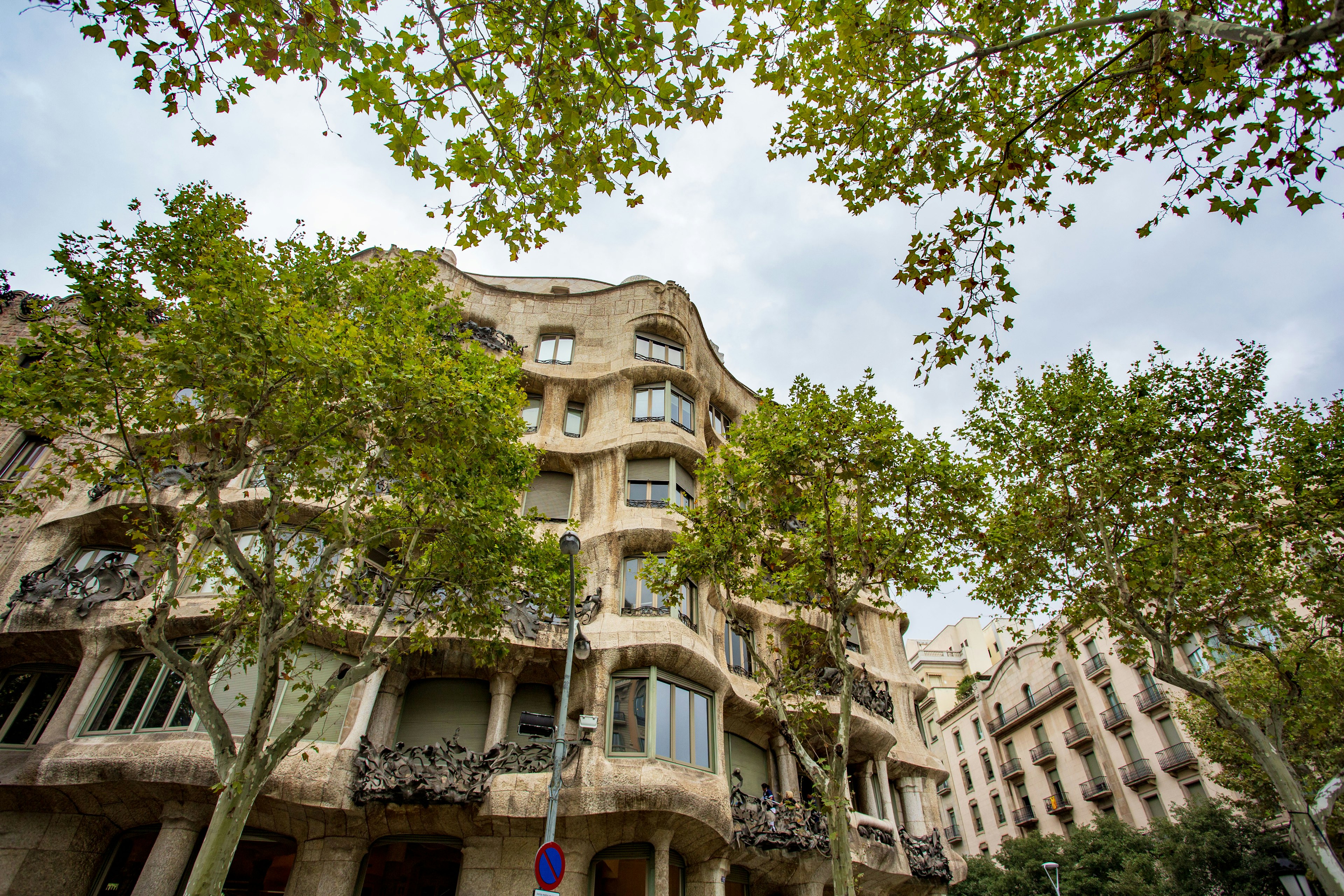 Exterior view of Casa Mila in Barcelona surrounded by green trees and a cloudy sky