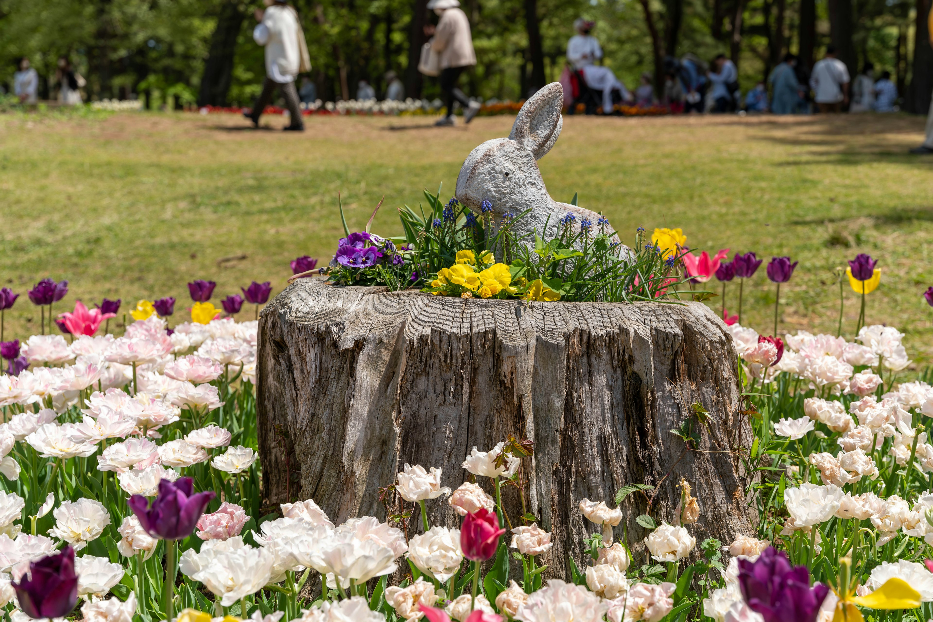 A stone rabbit sculpture on a tree stump surrounded by colorful flowers in a park