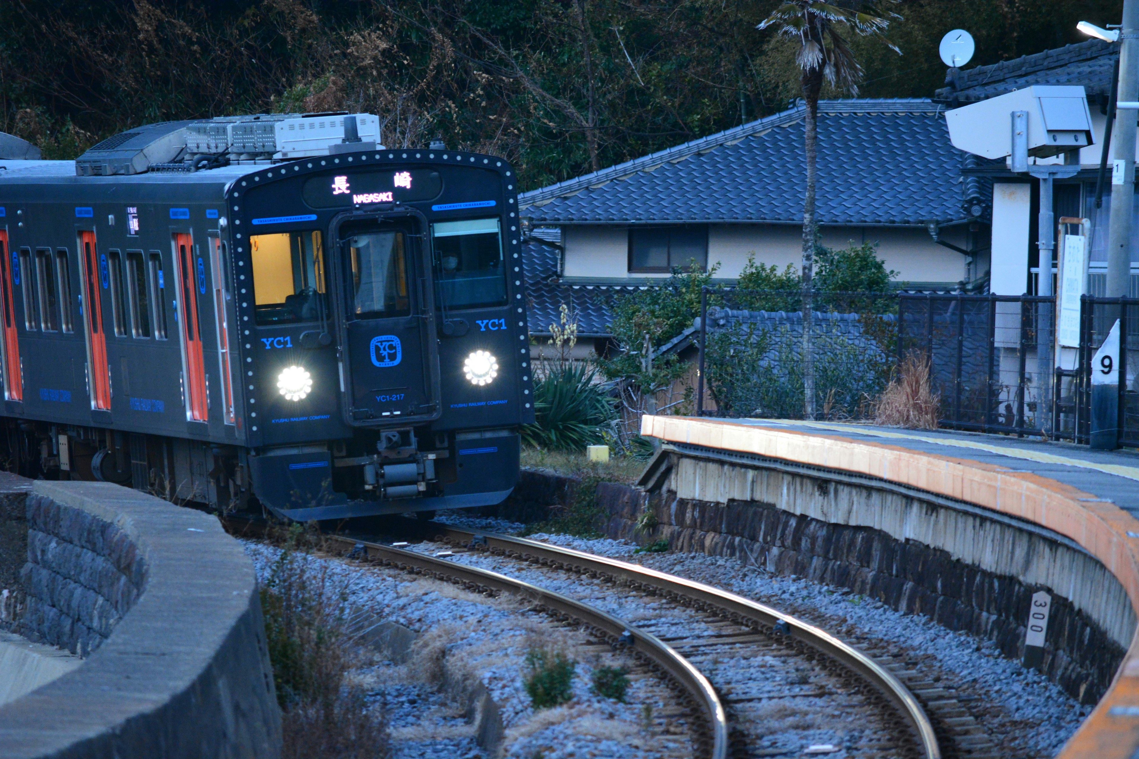 Tren azul acercándose por vías curvas cerca de casas tradicionales