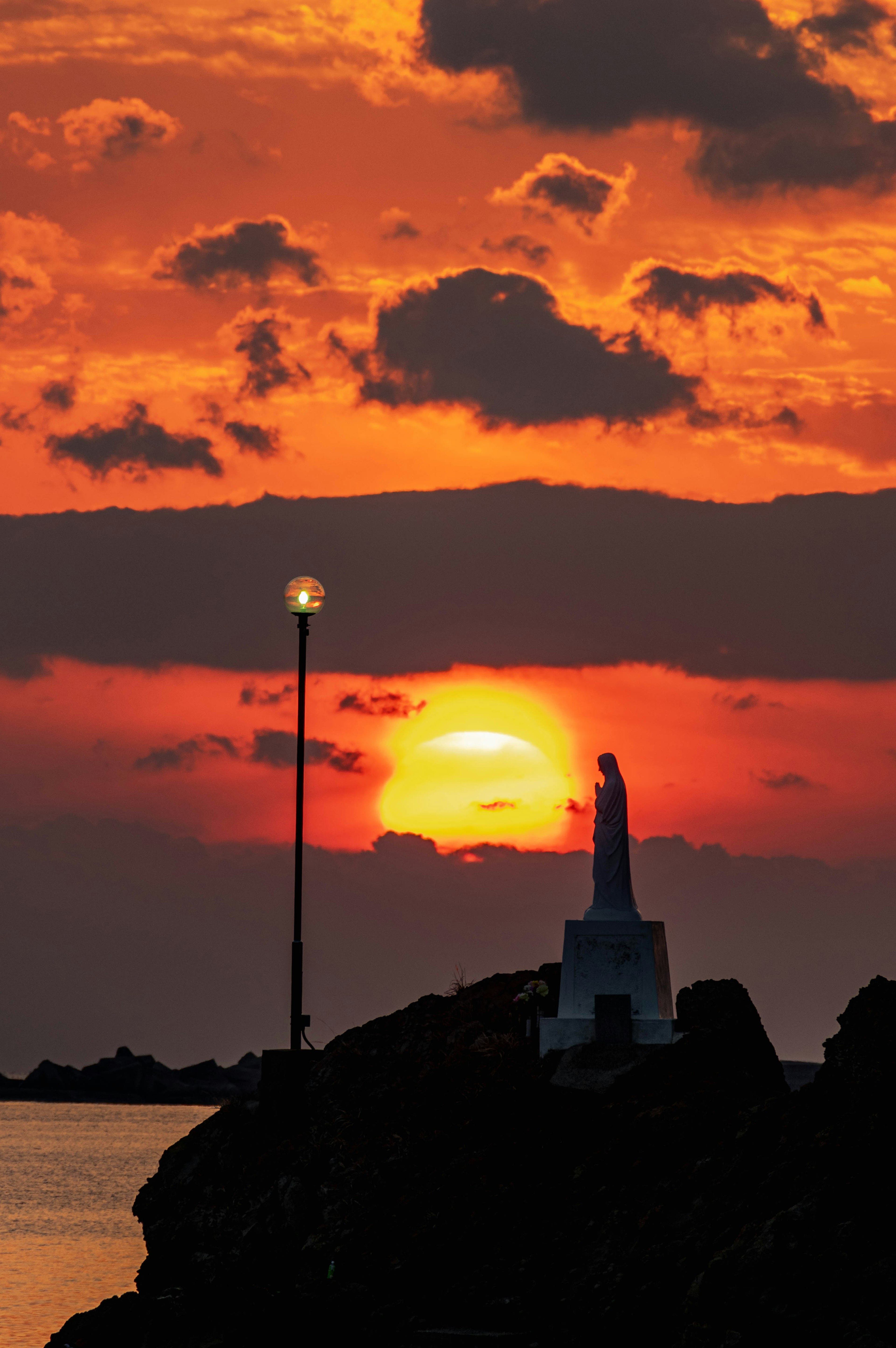 Silhouette einer Statue vor einem lebhaften Sonnenuntergang mit einer Straßenlaterne am Ufer