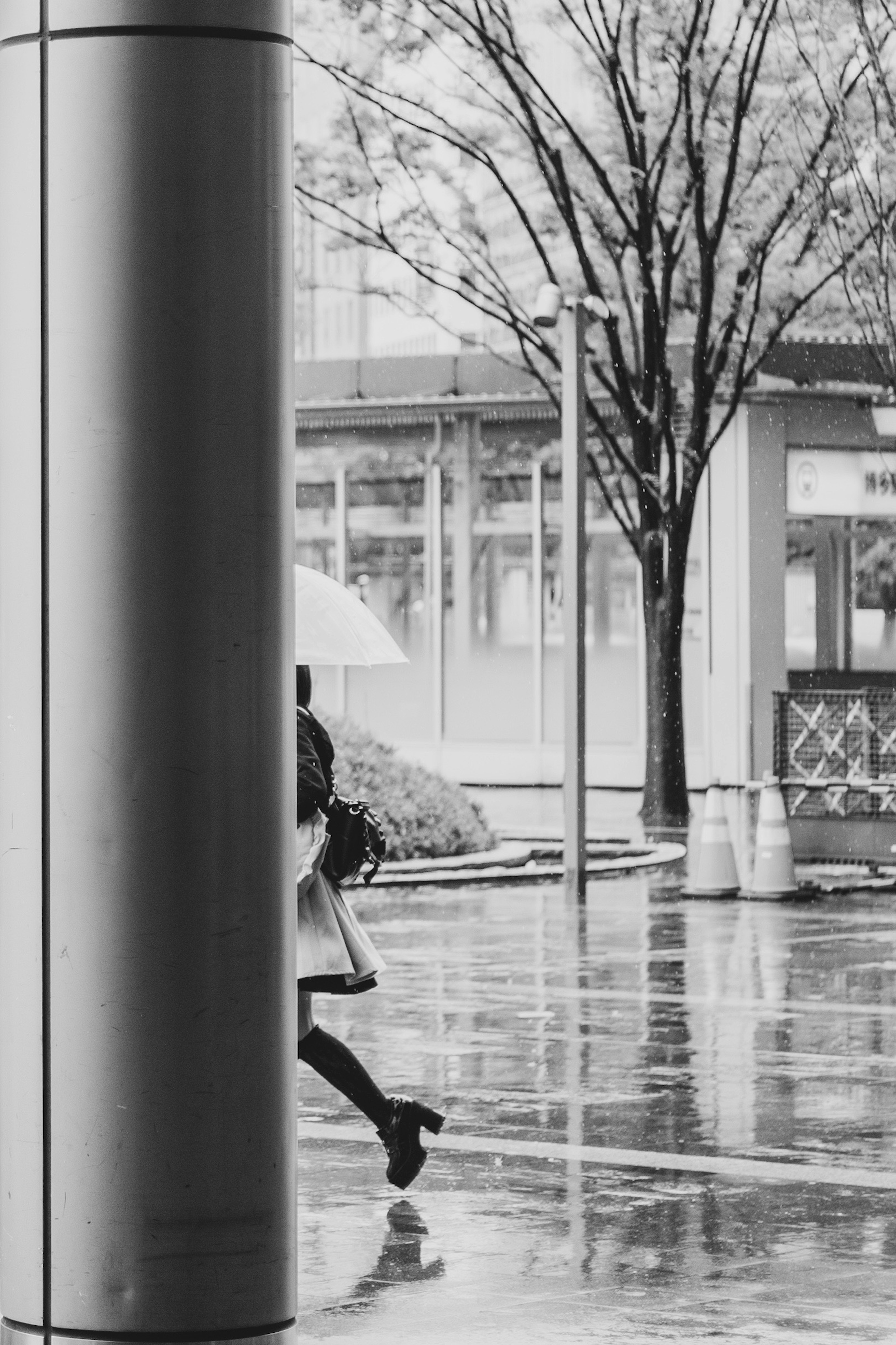 Black and white image of a person walking with an umbrella in the rain partially hidden behind a pillar