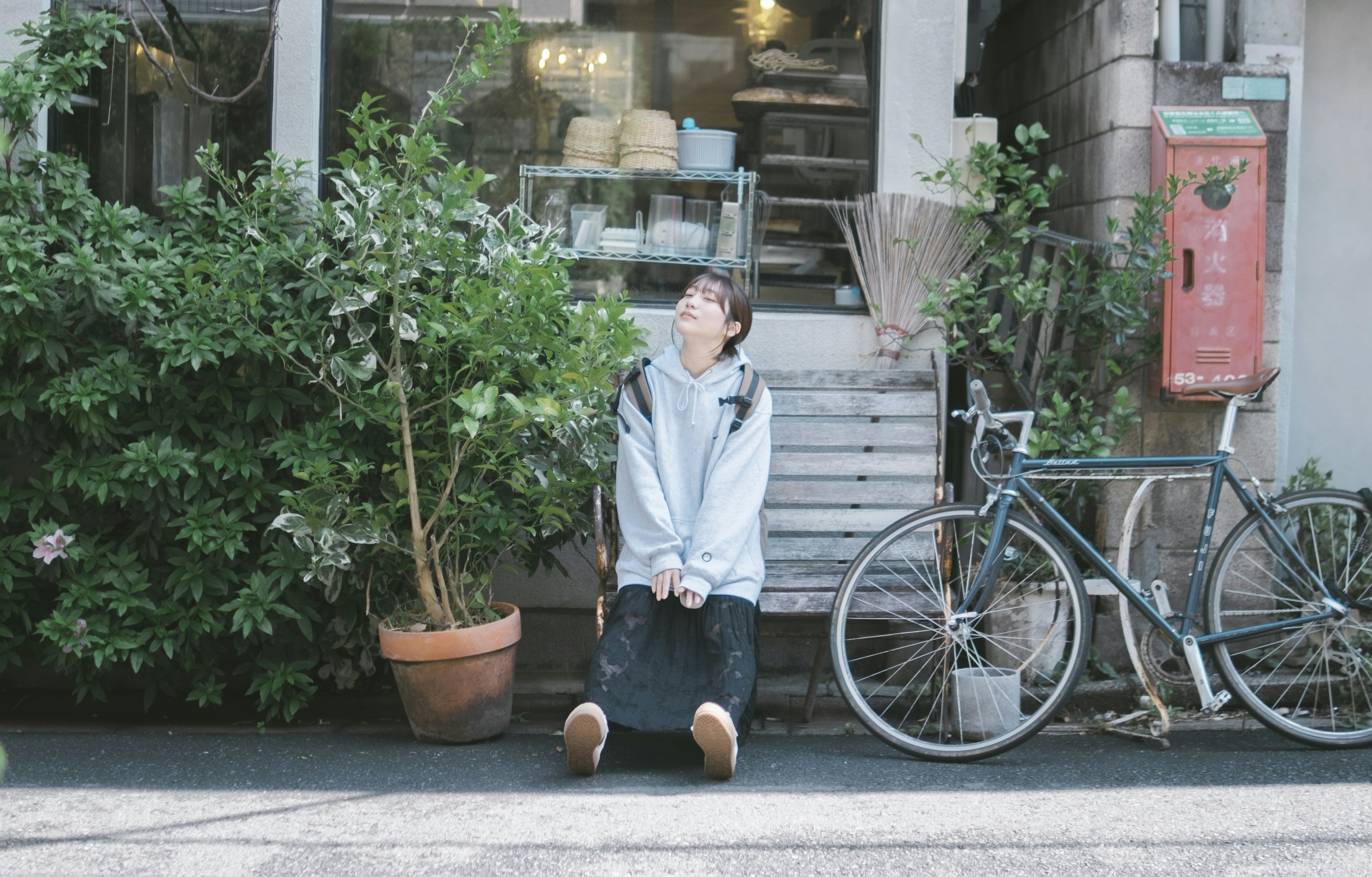 A young person sitting next to a bicycle outside a cafe featuring greenery and wooden steps