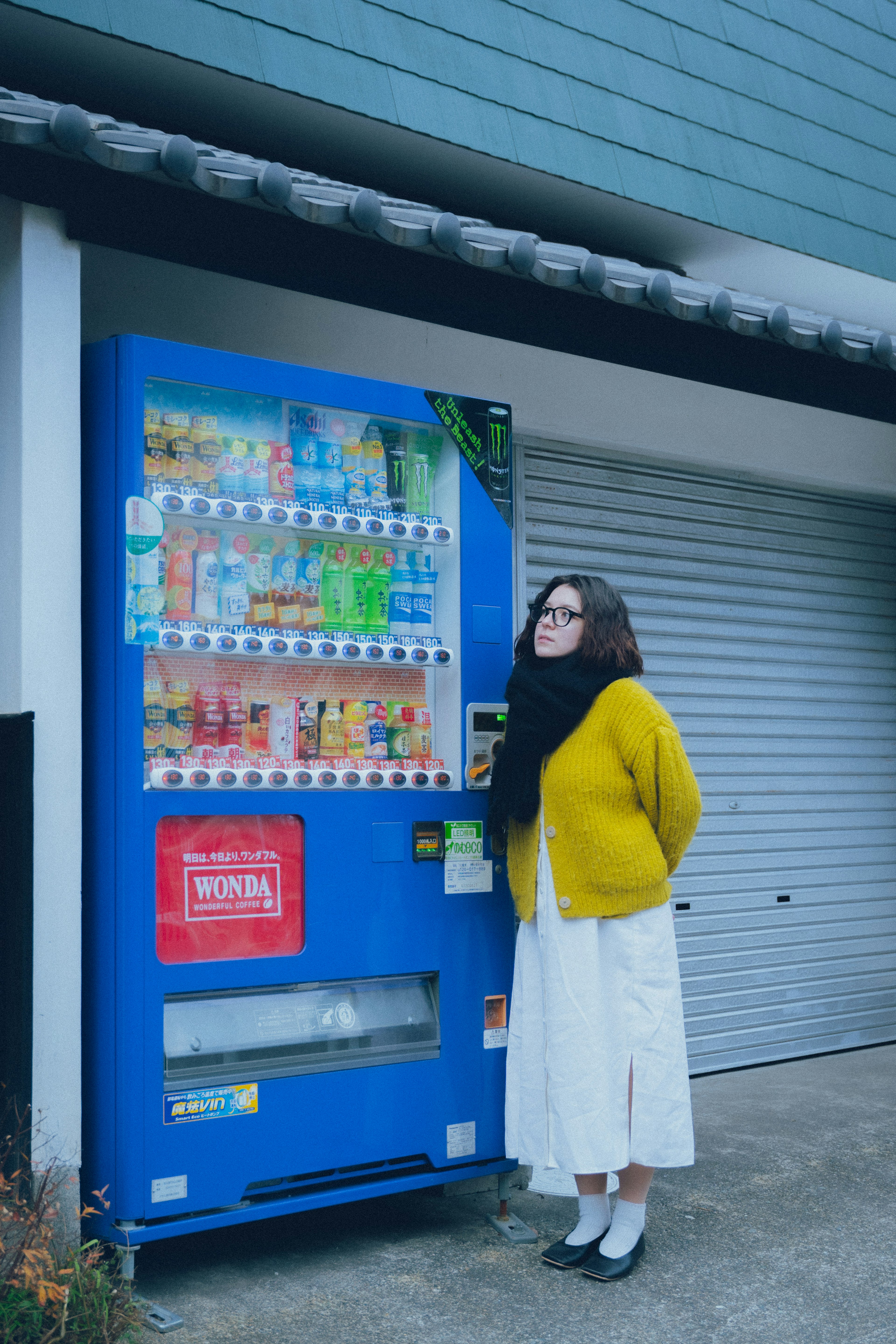 Donna in piedi davanti a una macchina automatica blu che indossa un maglione giallo e una gonna bianca lo sfondo presenta un ambiente residenziale