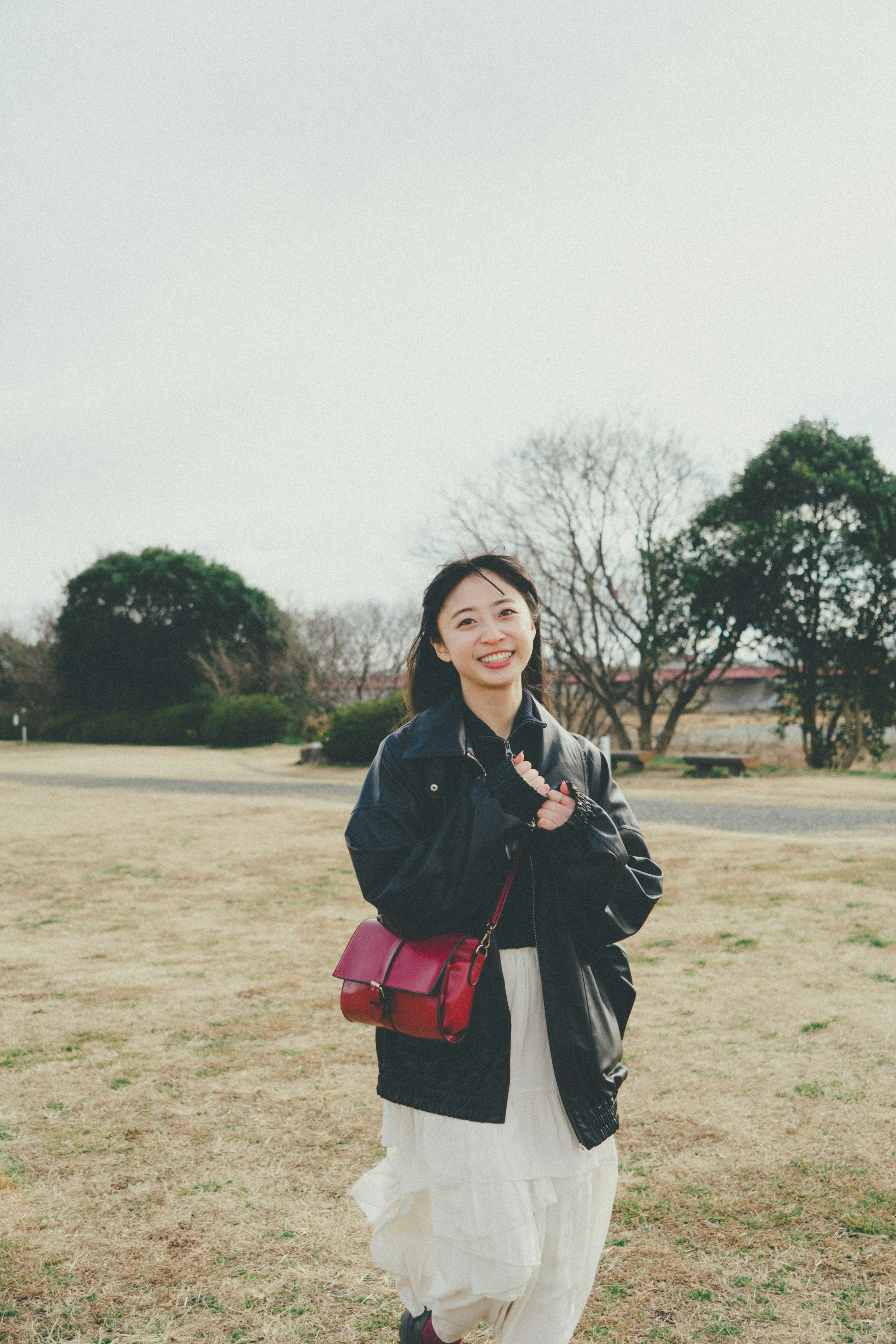 A woman walking in a park smiling wearing a black jacket and white dress carrying a red bag