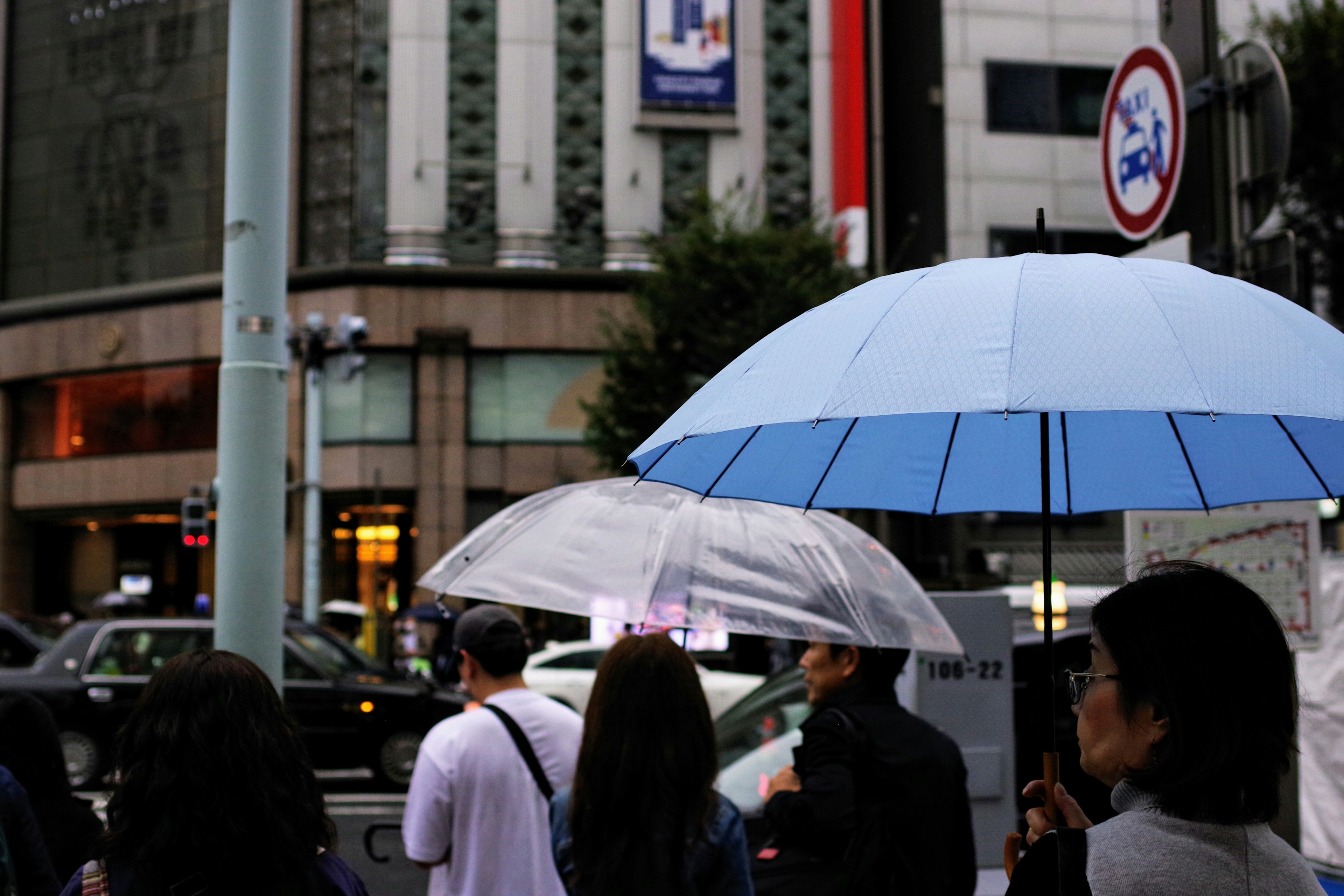 People holding umbrellas standing at a crosswalk with buildings and traffic signs in the background