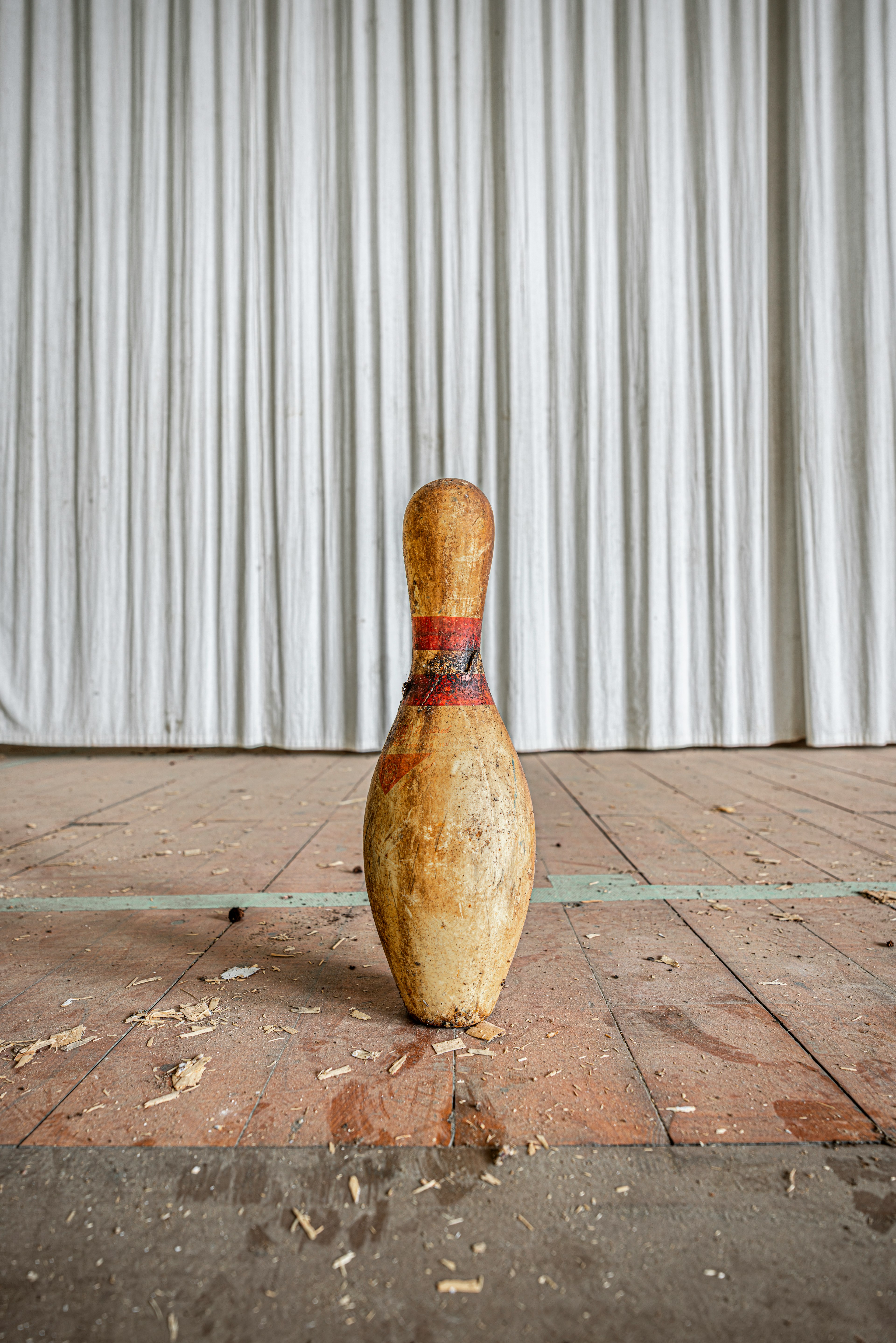 An old bowling pin standing on a wooden floor with a white curtain backdrop