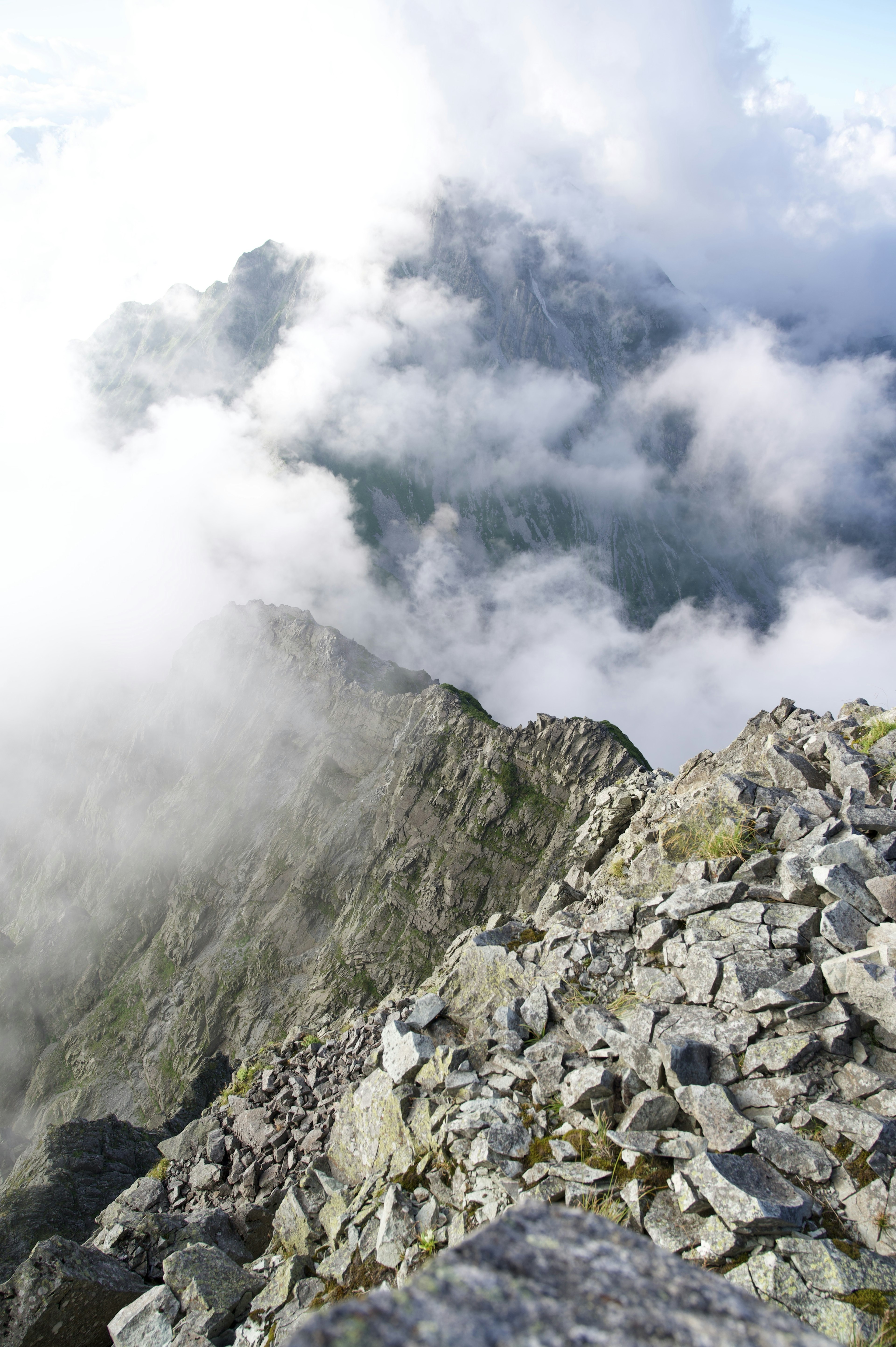 Blick von einem Berggipfel mit felsigem Gelände und Wolken, die die Berge bedecken