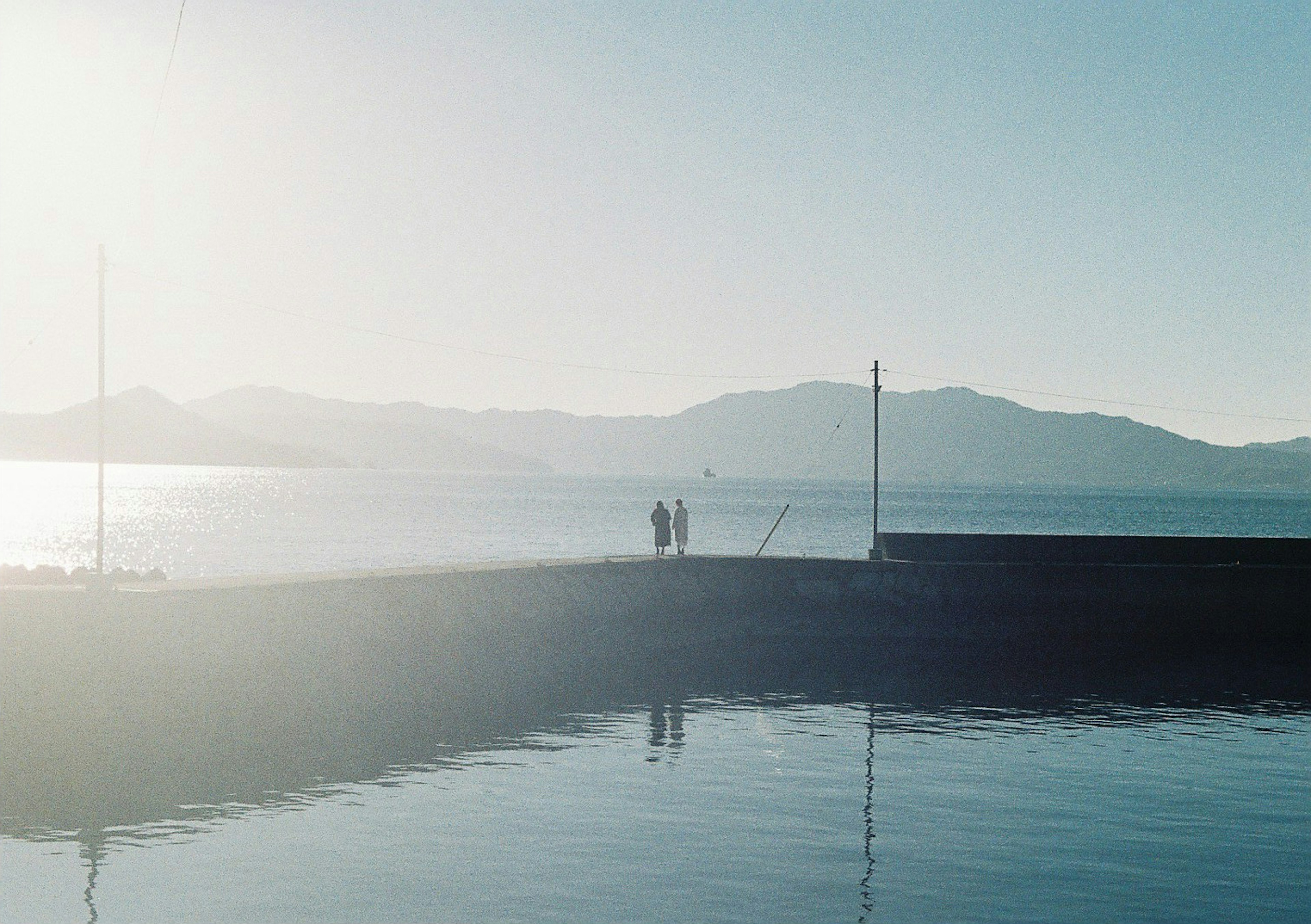 A serene harbor with a blue water surface and mountains in the background featuring two people standing