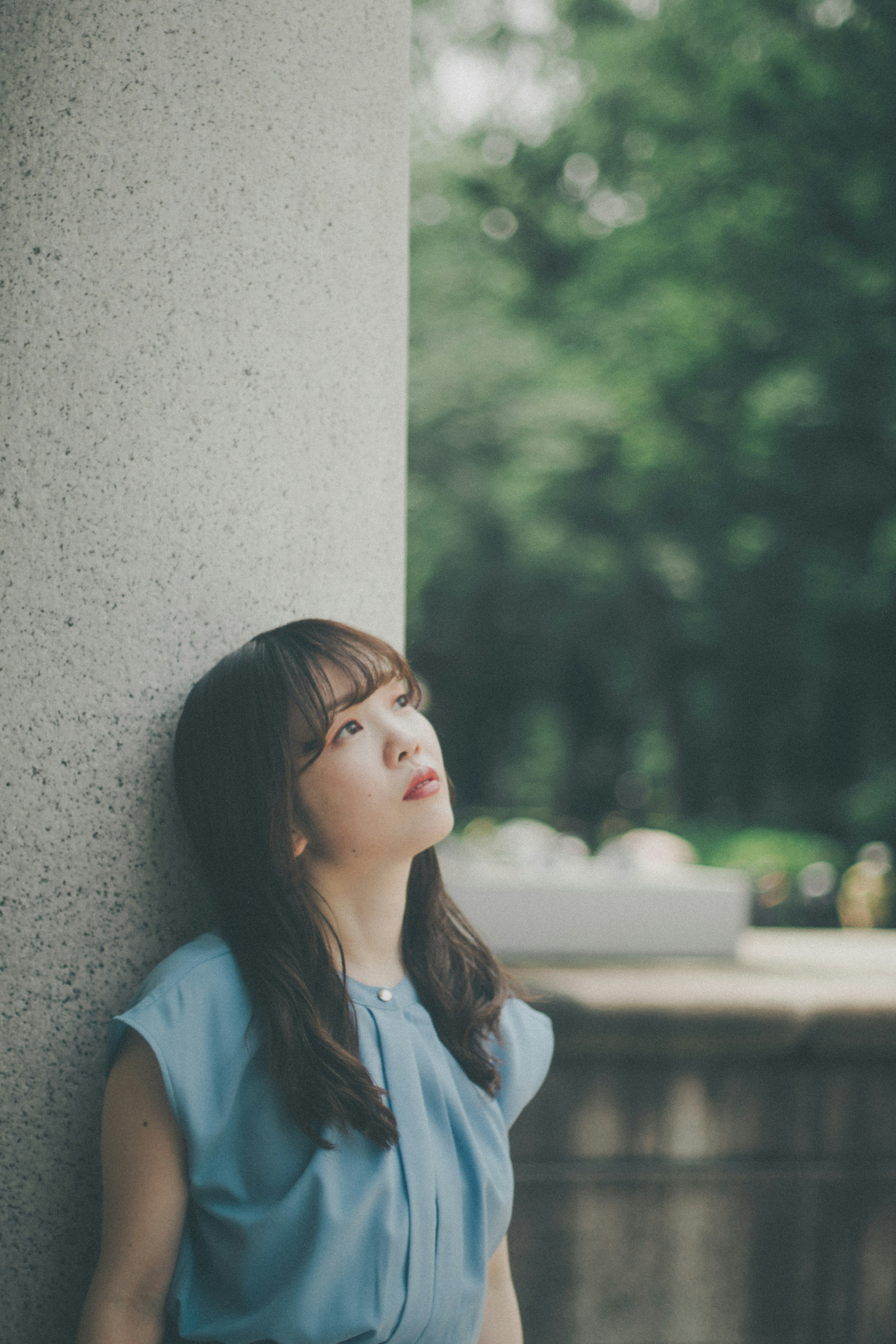 A woman in a light blue outfit leaning against a pillar looking up thoughtfully