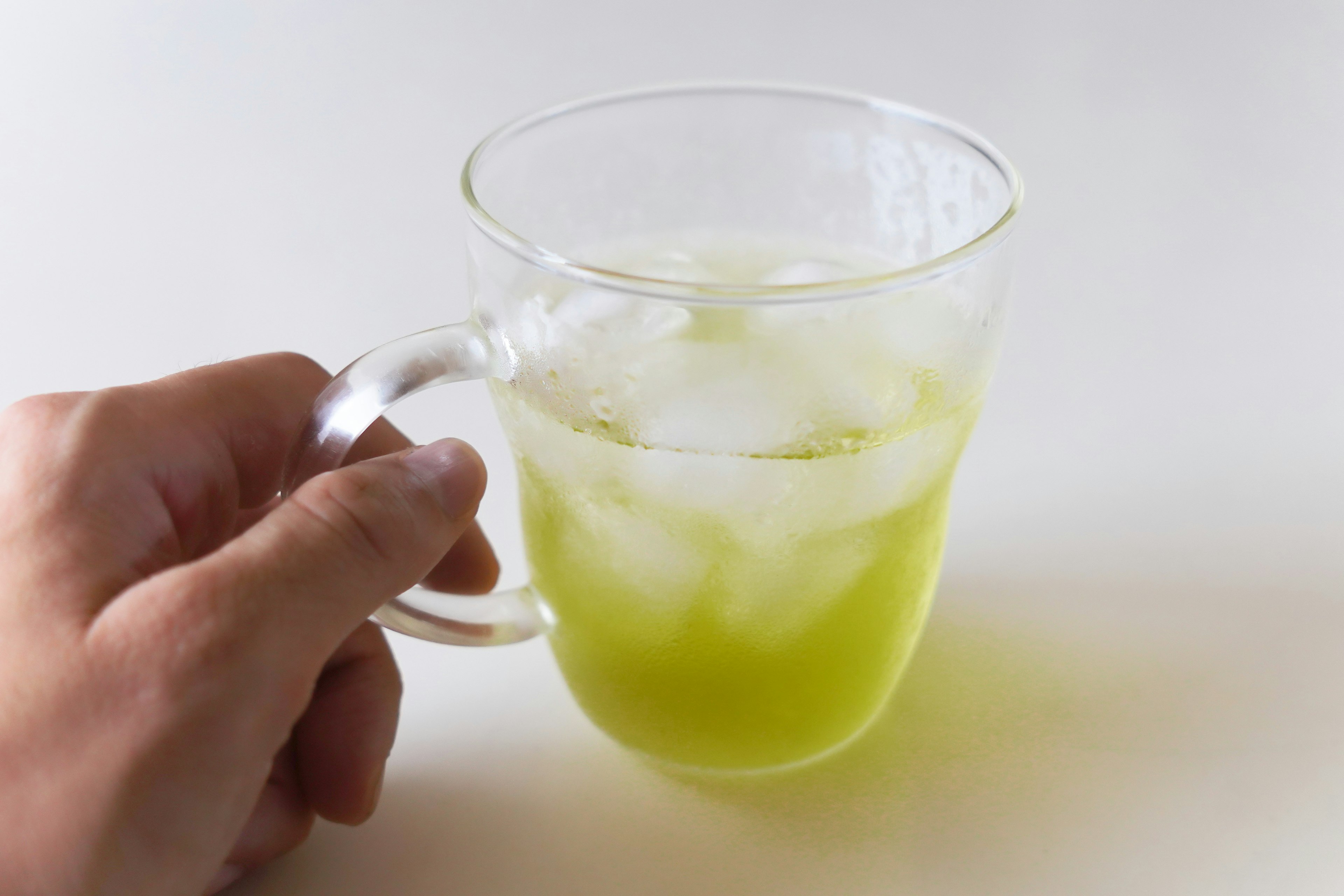 A hand holding a clear glass mug containing green drink with ice