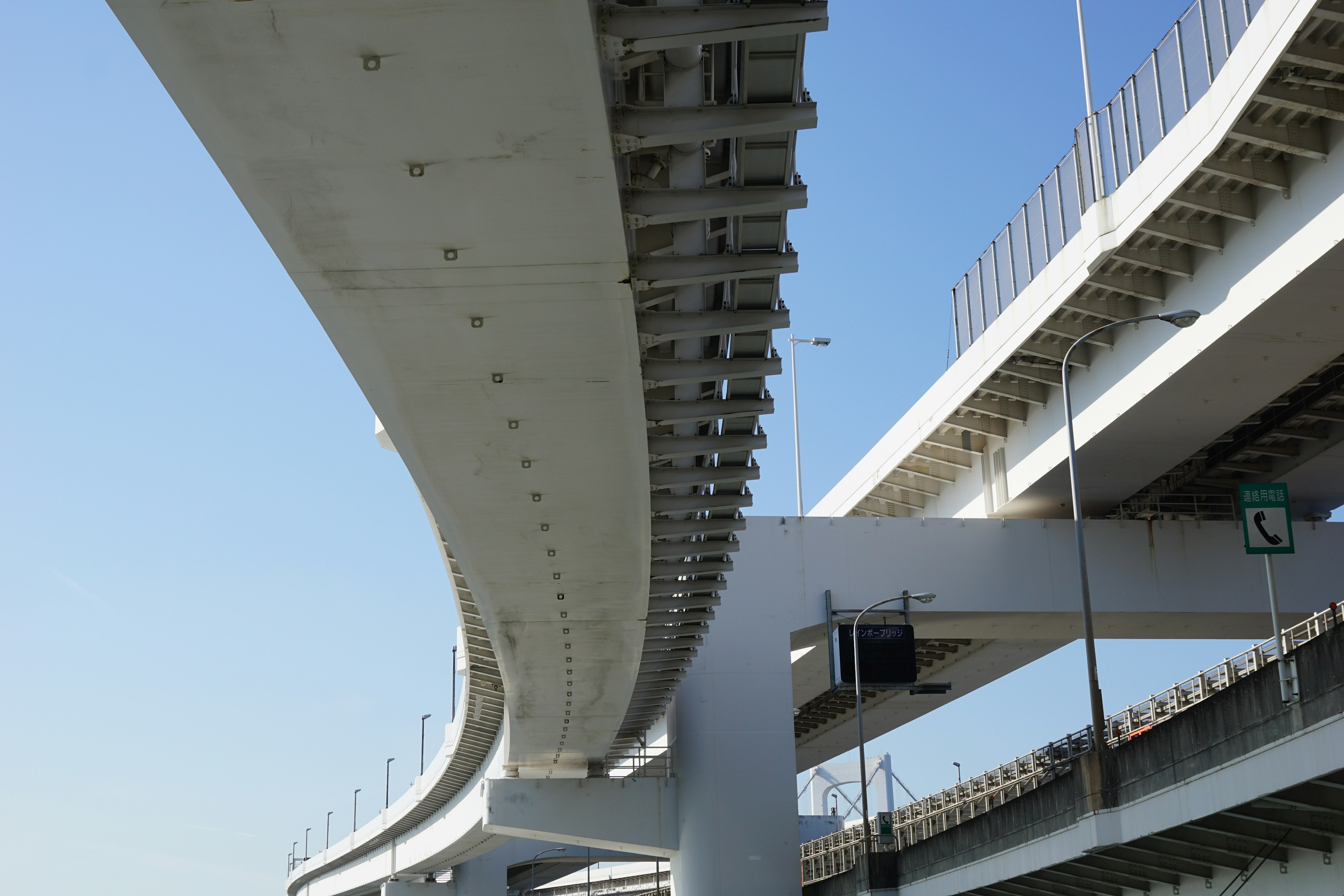 Foto jembatan layang dilihat dari bawah dengan struktur beton melengkung di bawah langit biru yang cerah