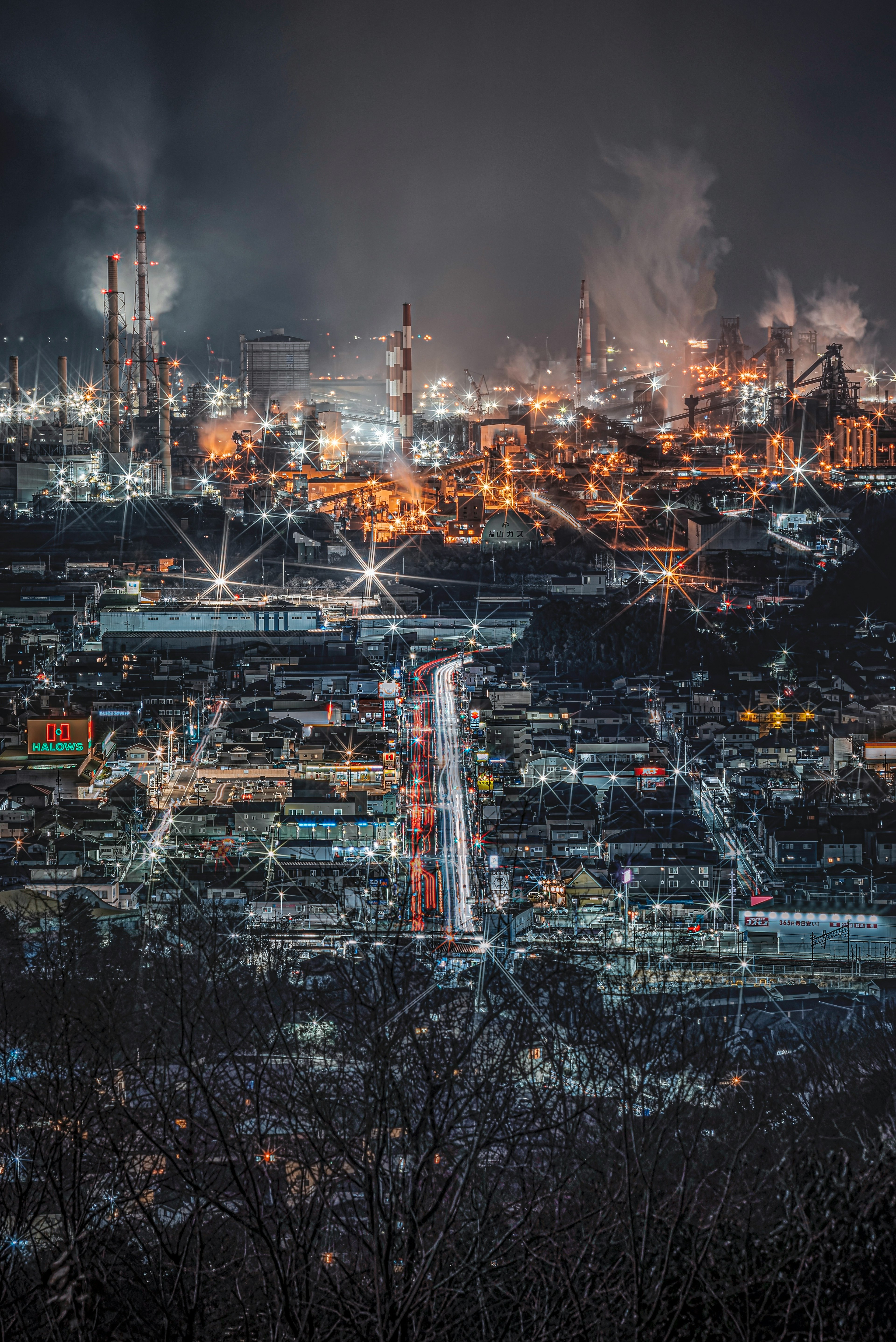 Vista notturna di un'area industriale con fabbriche e camini illuminati dalle luci della città