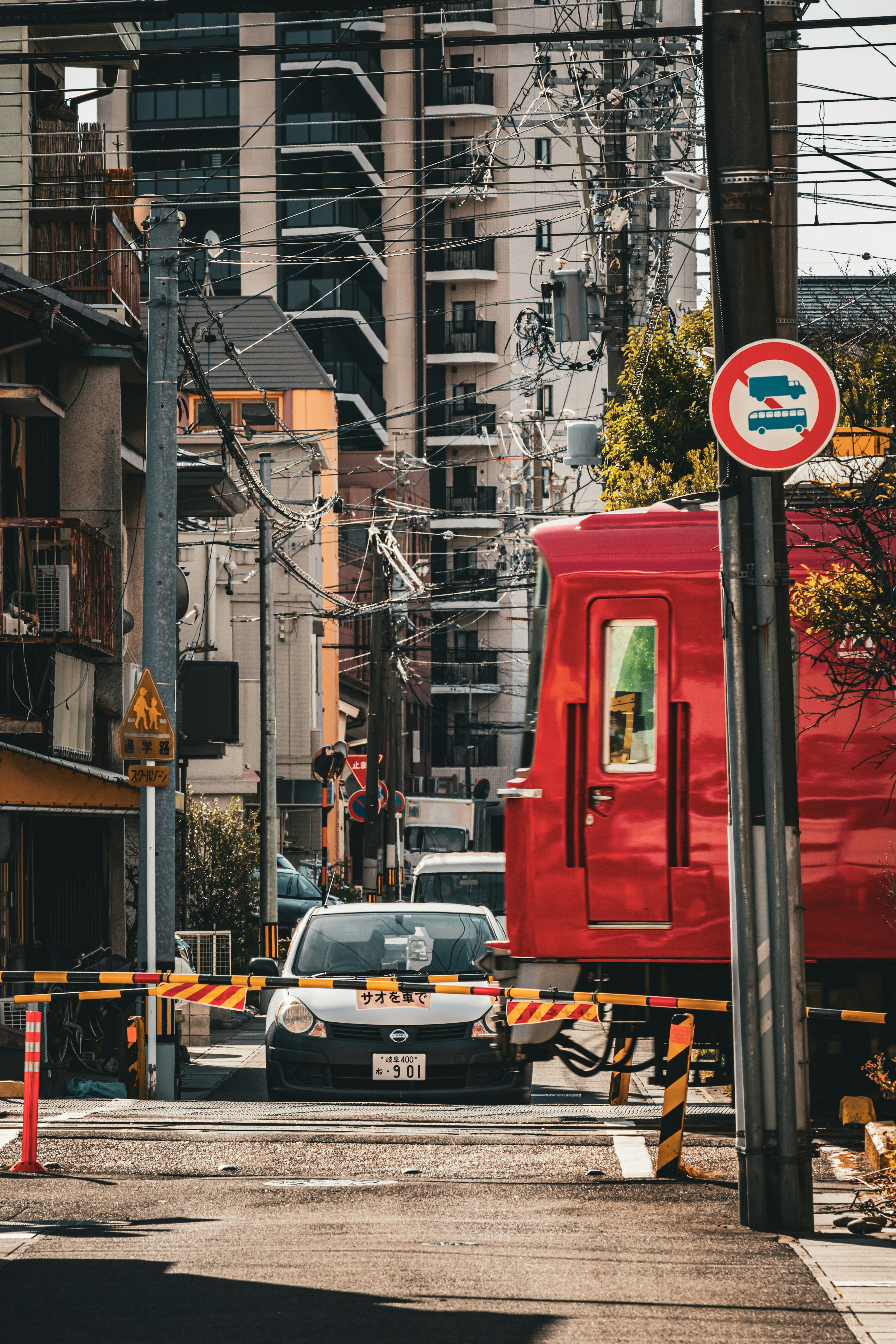 A red train passing through an urban street with high-rise buildings and a traffic sign visible