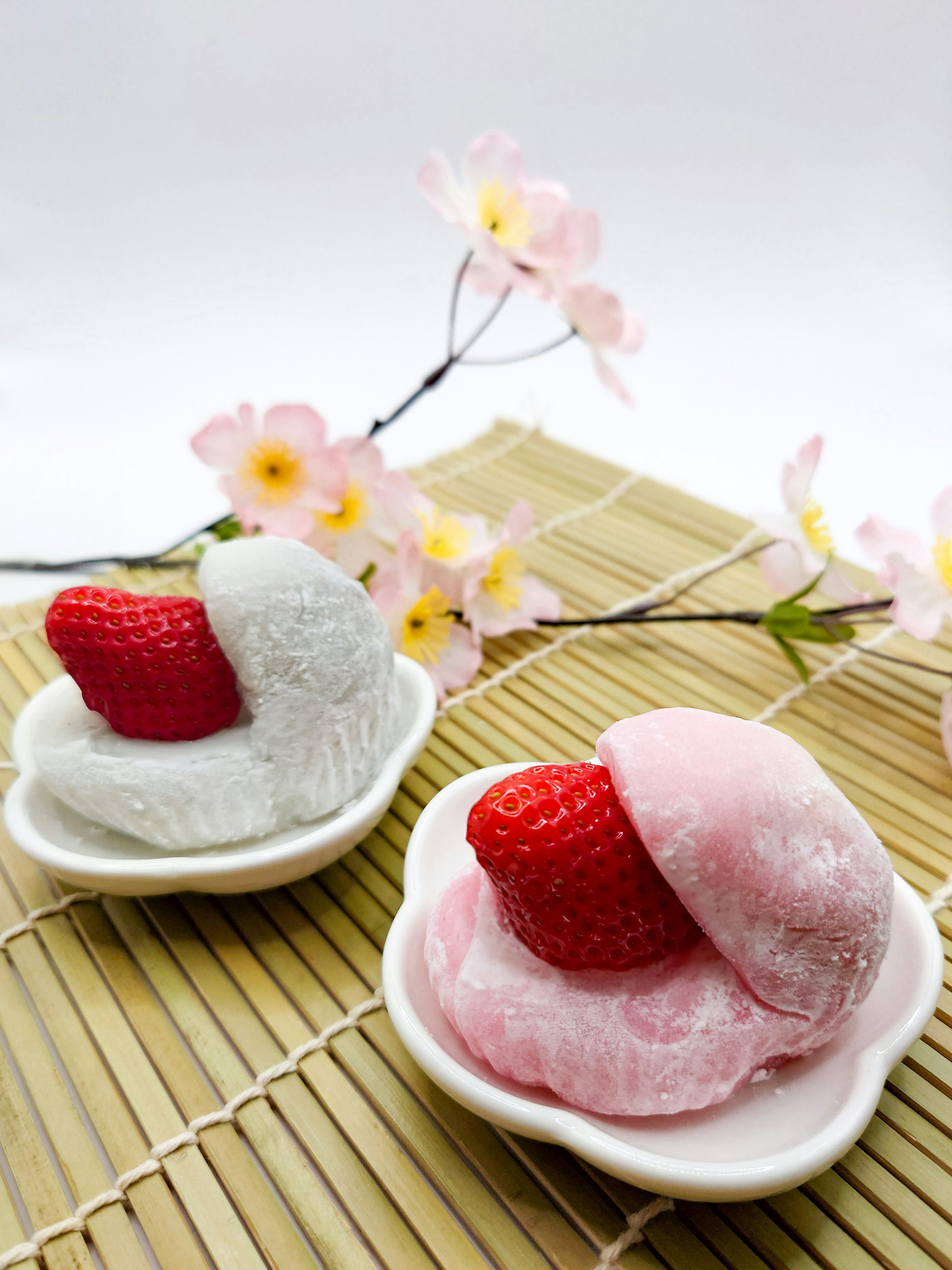 Dos daifuku de fresa en blanco y rosa sobre una estera de bambú con flores de cerezo